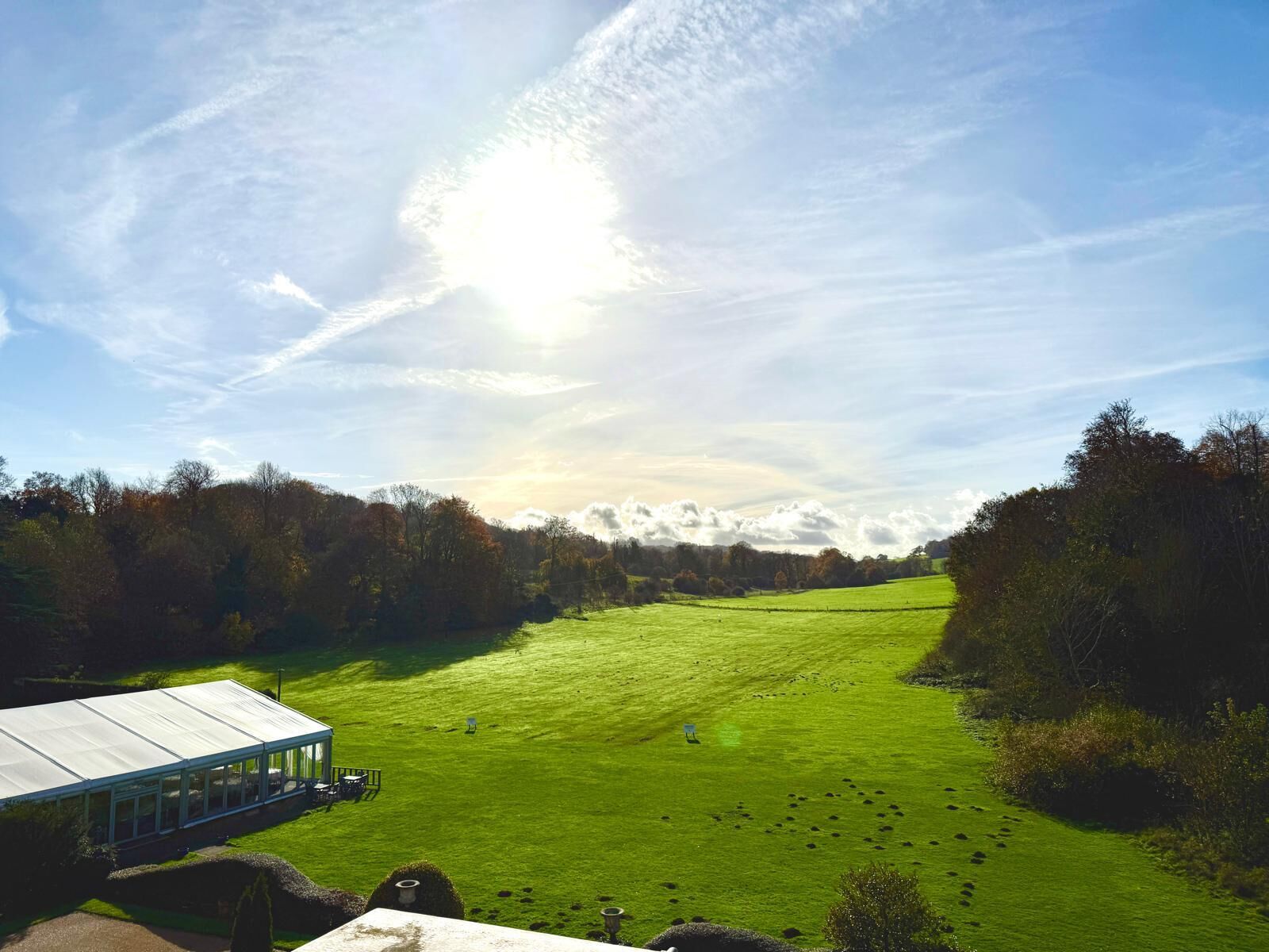 A green field with a glass-roofed structure, likely a greenhouse or conservatory, surrounded by trees.