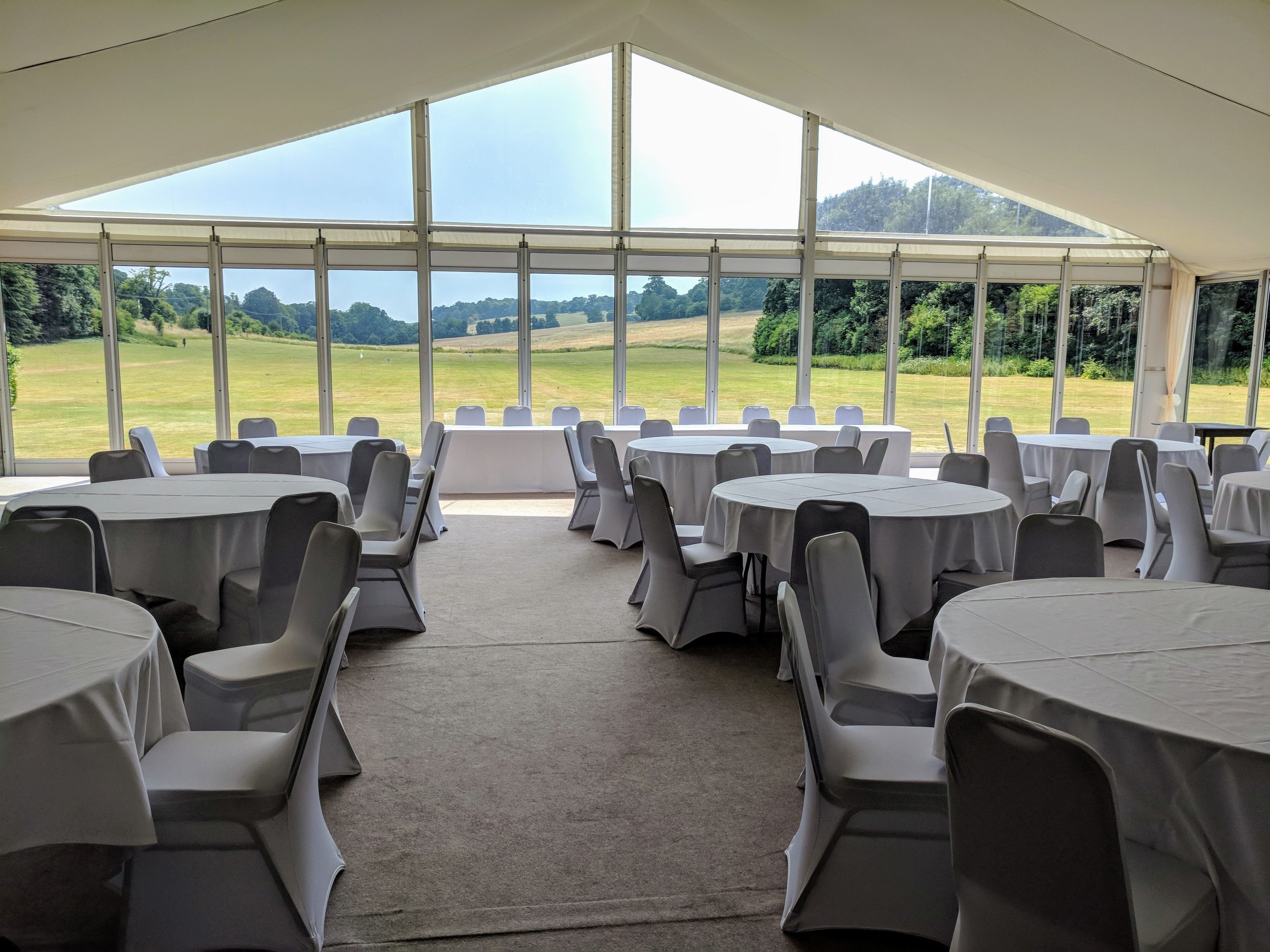 A tented event space with round tables and chairs, large windows offering a view of a grassy field.