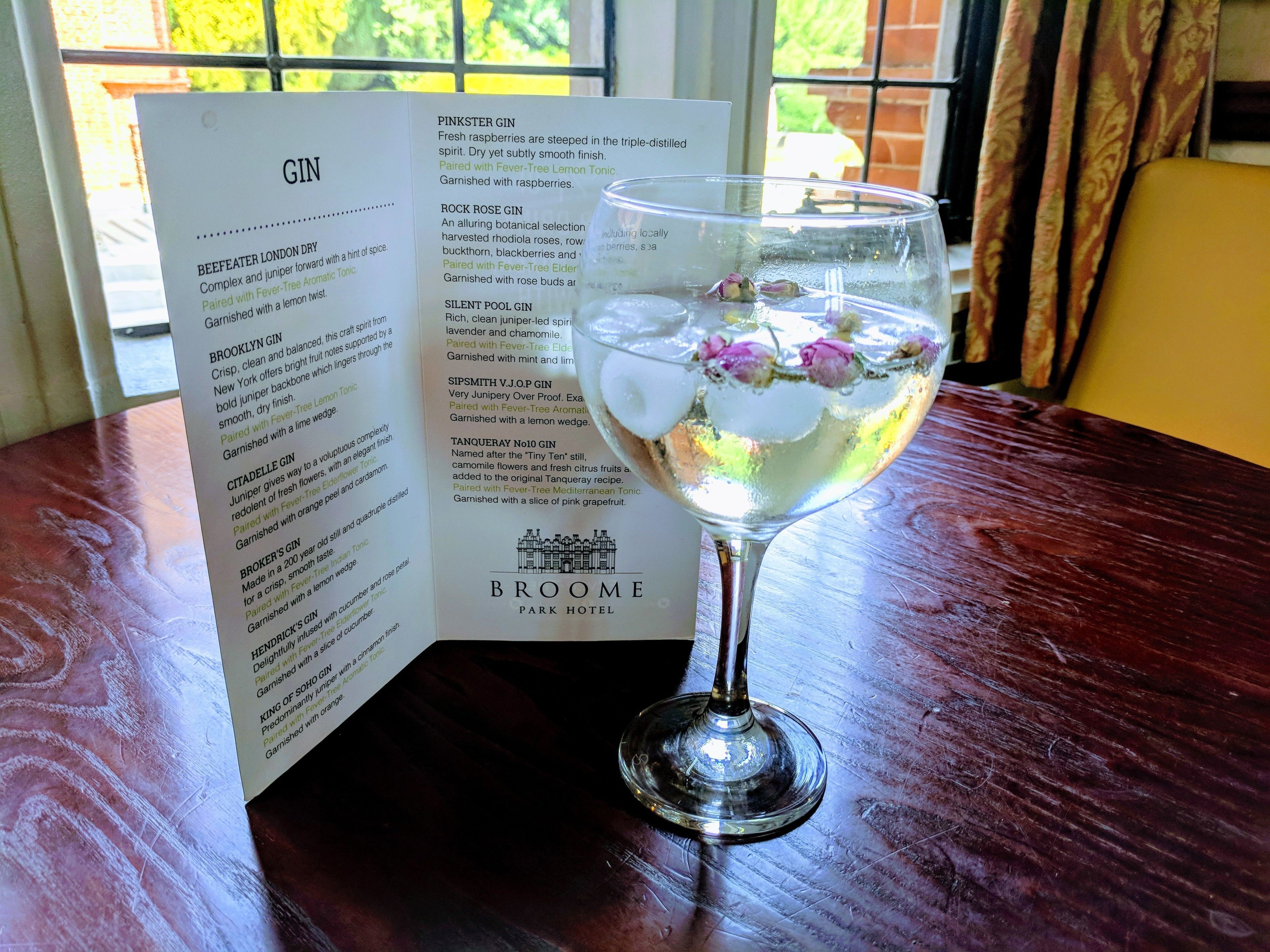 A menu with various gin selections and a glass of gin with ice and flowers on a wooden table.