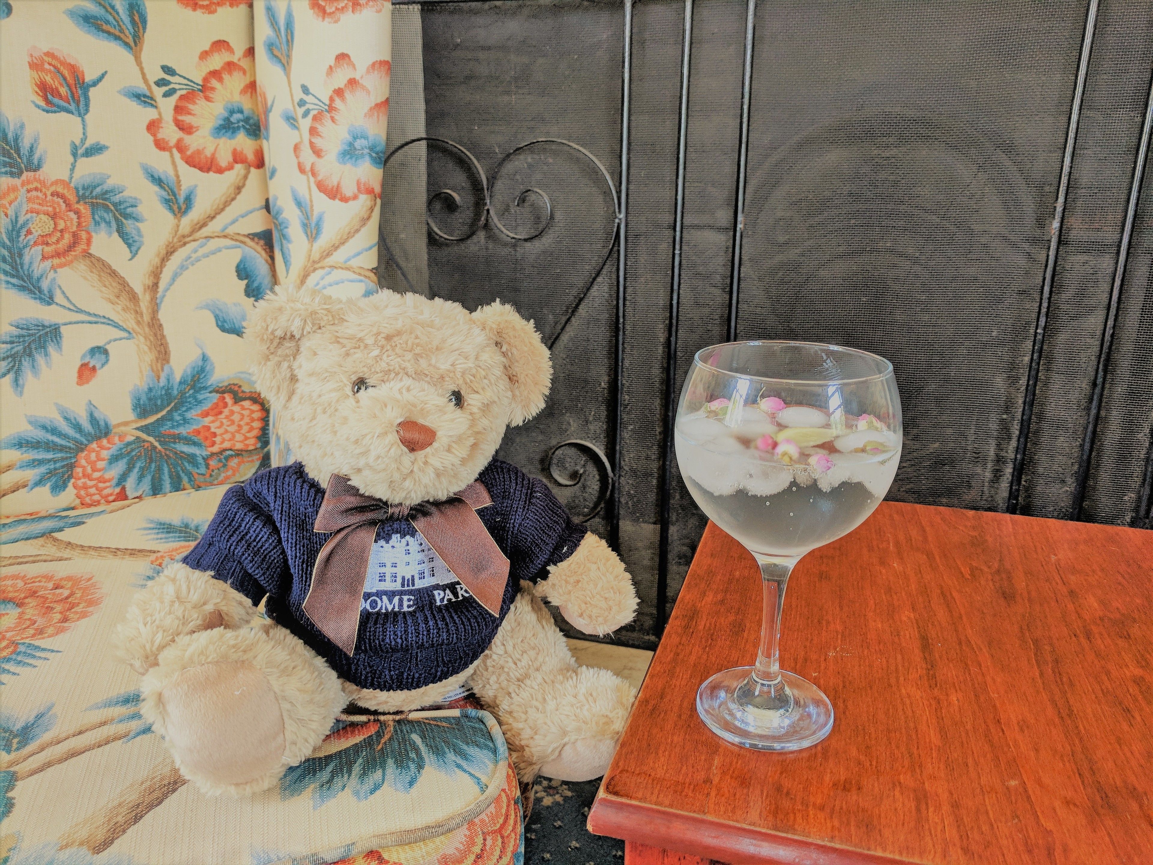 Un oso de peluche sobre una silla con estampado floral, junto a una mesa de madera con un vaso de vidrio transparente que contiene una bebida.