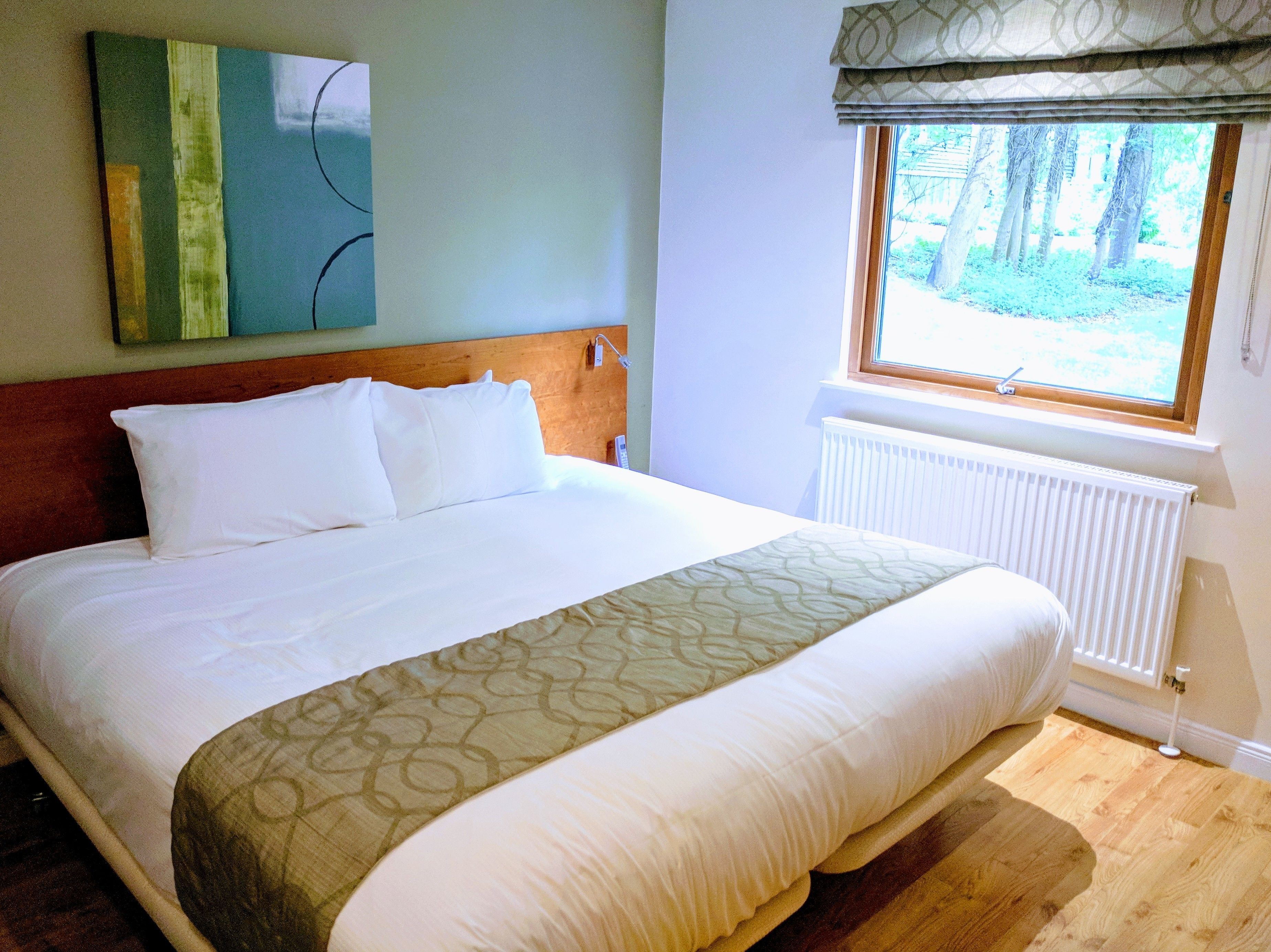 A neatly made bed with white linens and a patterned throw, a wooden headboard, a window with a view of trees, and a radiator.