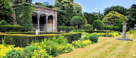 Un jardín con una estructura de ladrillo que presenta arcos, rodeado de setos cuidados y plantas con flores.