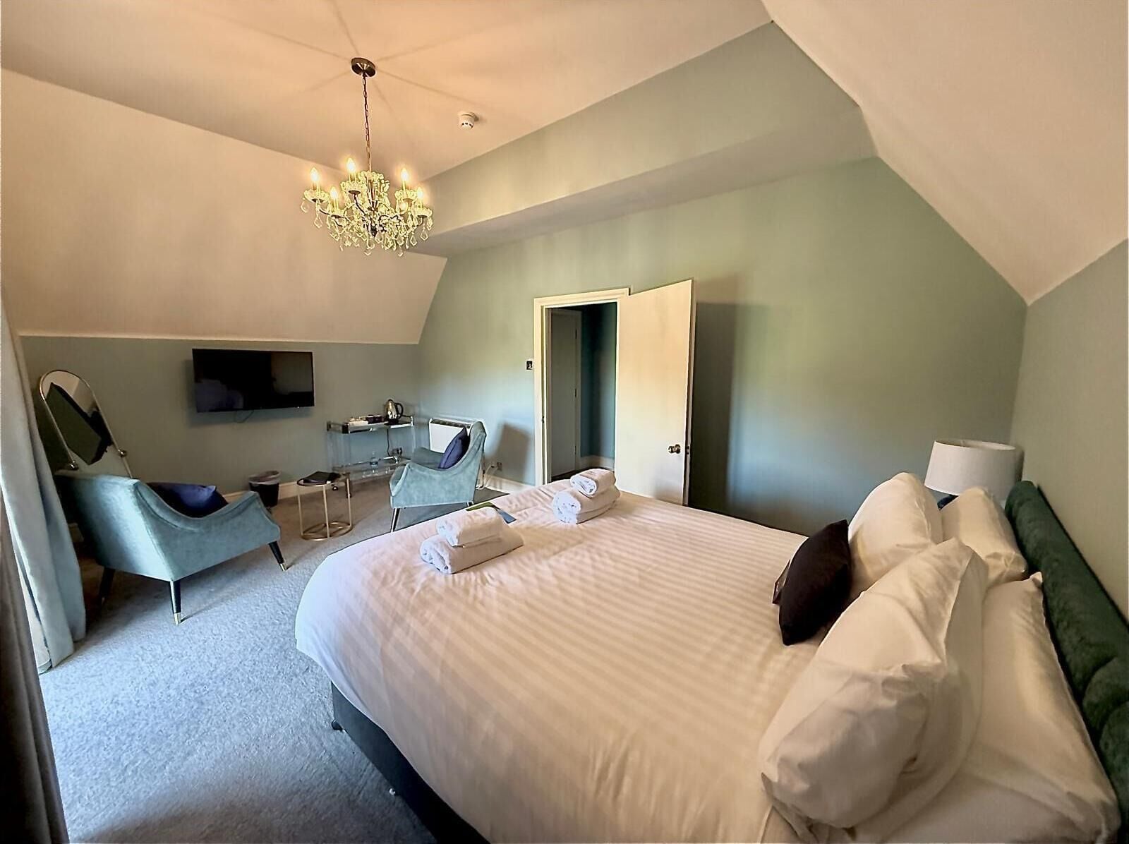 A bedroom with a bed, a chair, a desk, a mirror, a TV, and a chandelier.