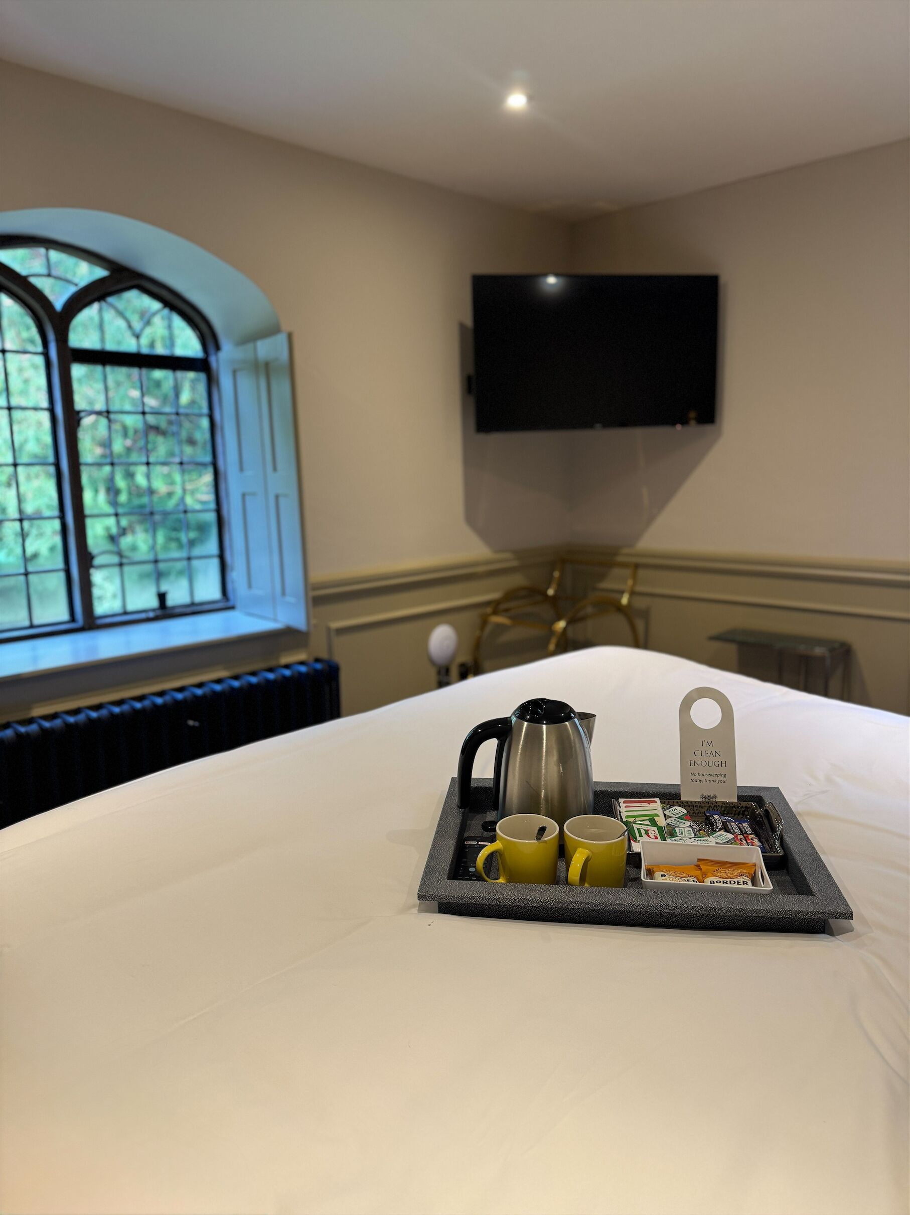 Quarto de hotel com uma cama, um suporte com chaleira, duas canecas e um aviso escrito 'Limpo o suficiente'.
