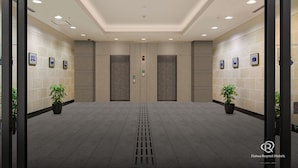 En moderne hotellobby med et rent flisegulv, en central elevator og krukkeplanter på hver side.