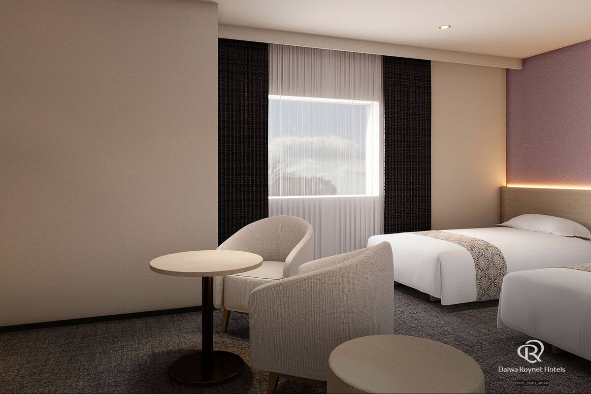 Una habitación de hotel con cama, dos sillas, una mesita, una ventana con cortinas y una lámpara fijada a la pared.