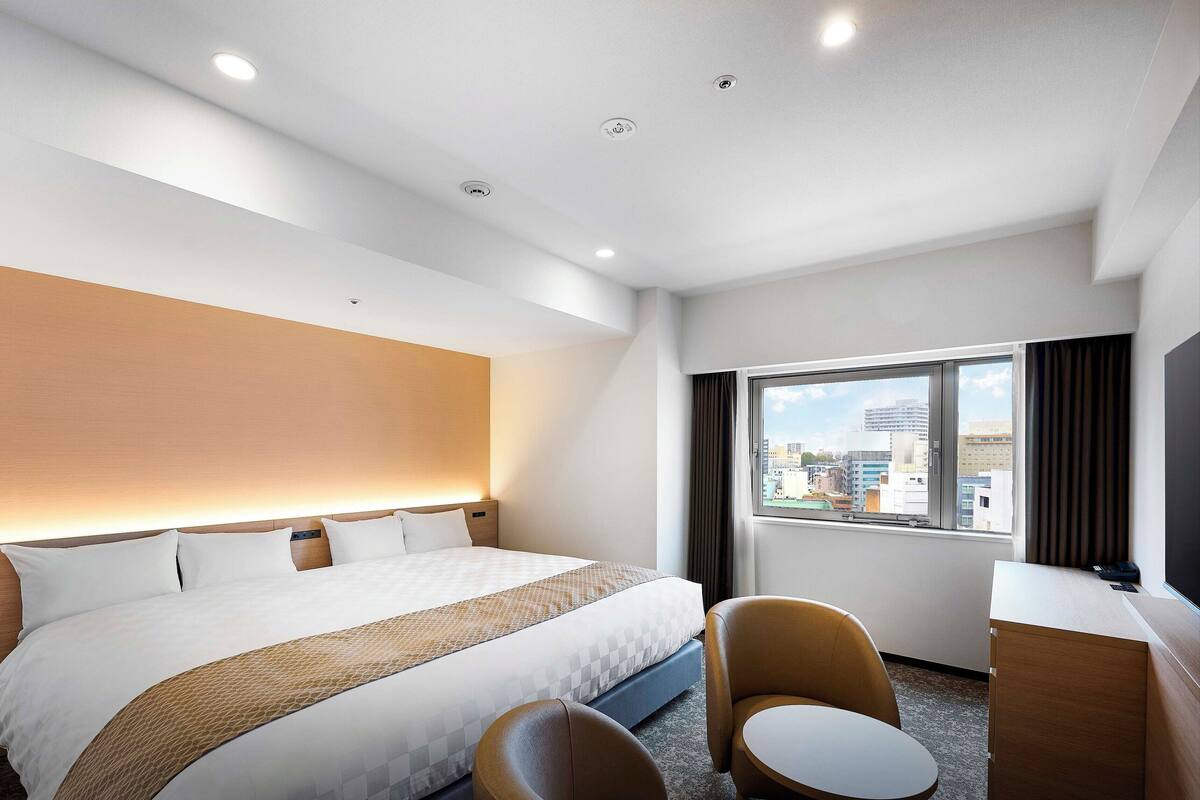 Habitación de hotel con una cama grande, un escritorio, una silla y vistas a la ciudad desde la ventana.