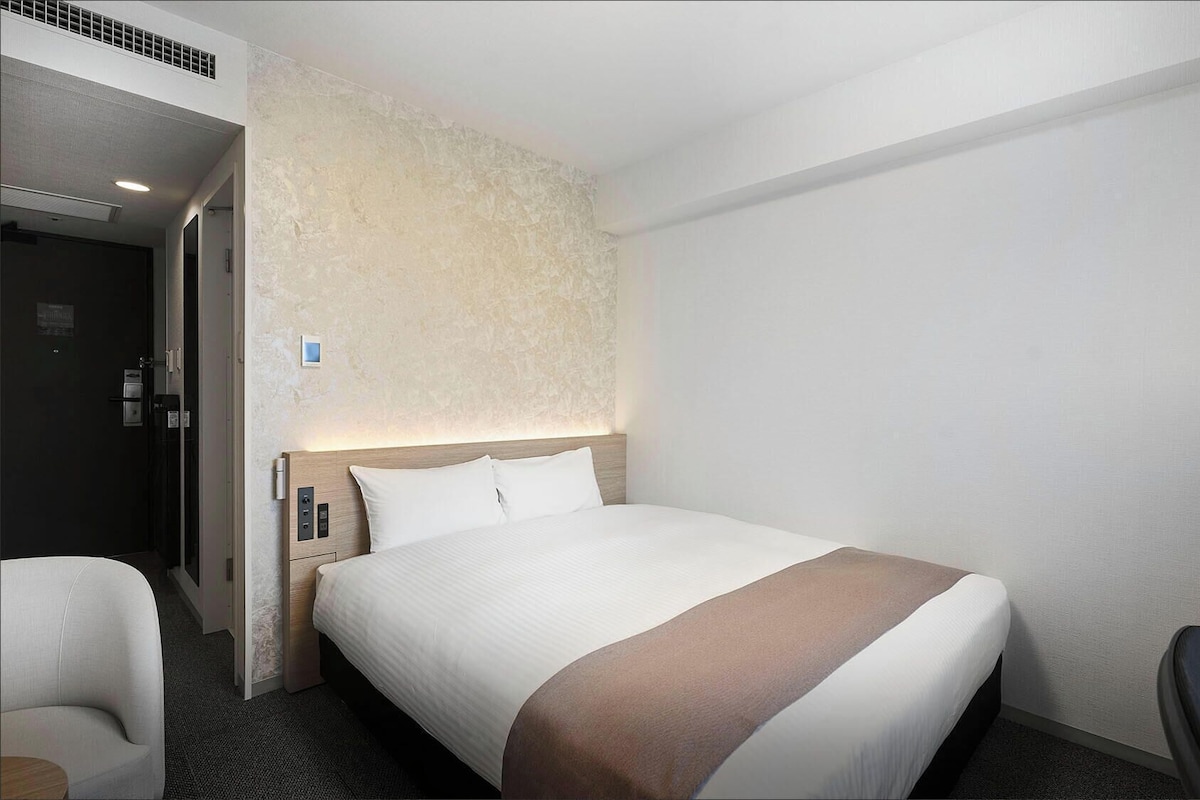 Una habitación de hotel con cama, una silla, una mesita de noche y un panel de control en la pared.