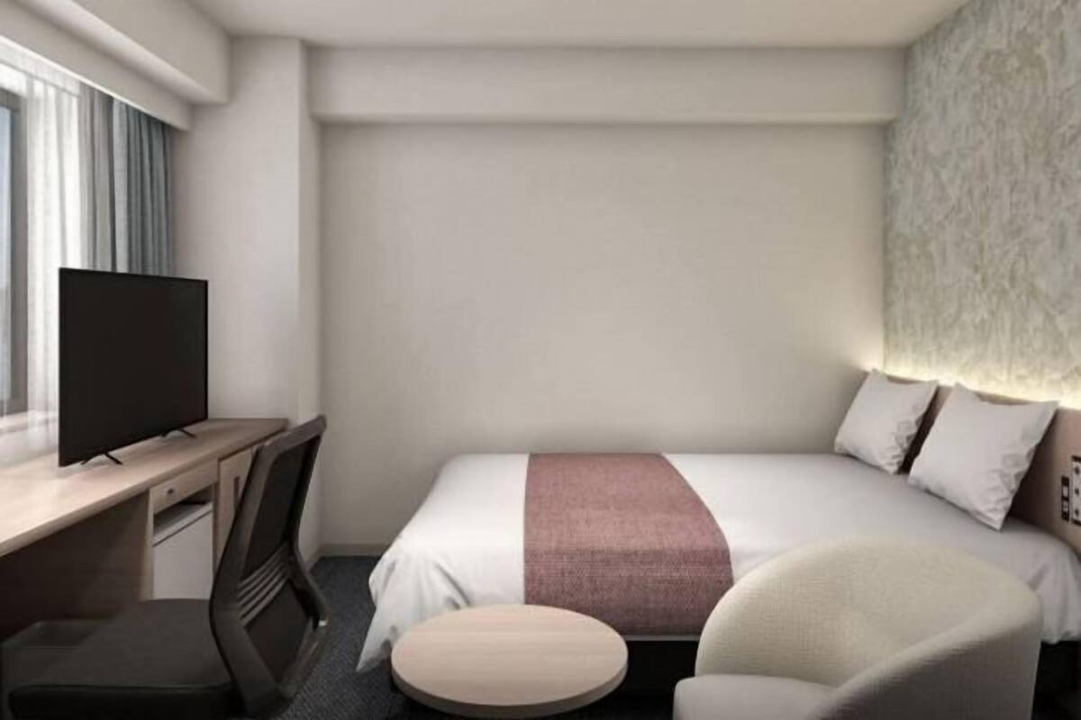 A hotel room with a bed, a desk with a television, a chair, and a round ottoman.