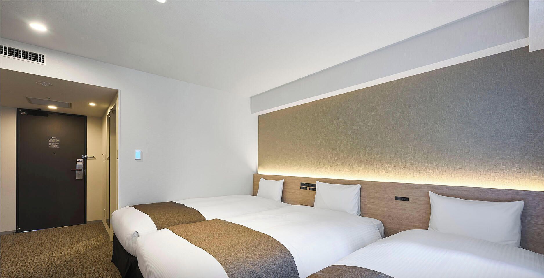 Une chambre d’hôtel avec deux lits, une porte sombre et un mur de couleur claire orné d’un motif texturé.