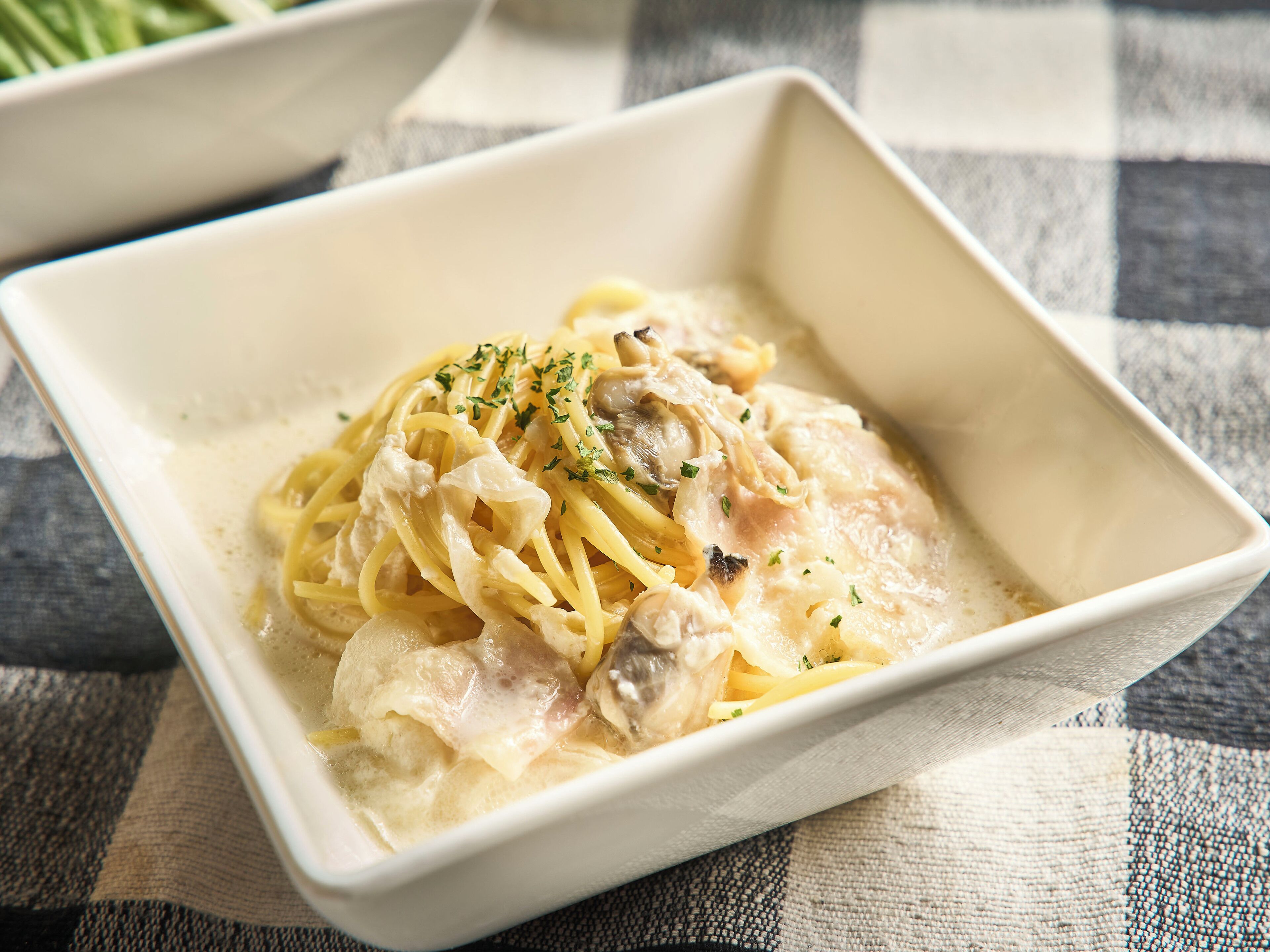 A square white bowl containing a creamy pasta dish with mushrooms and herbs, placed on a checkered cloth.