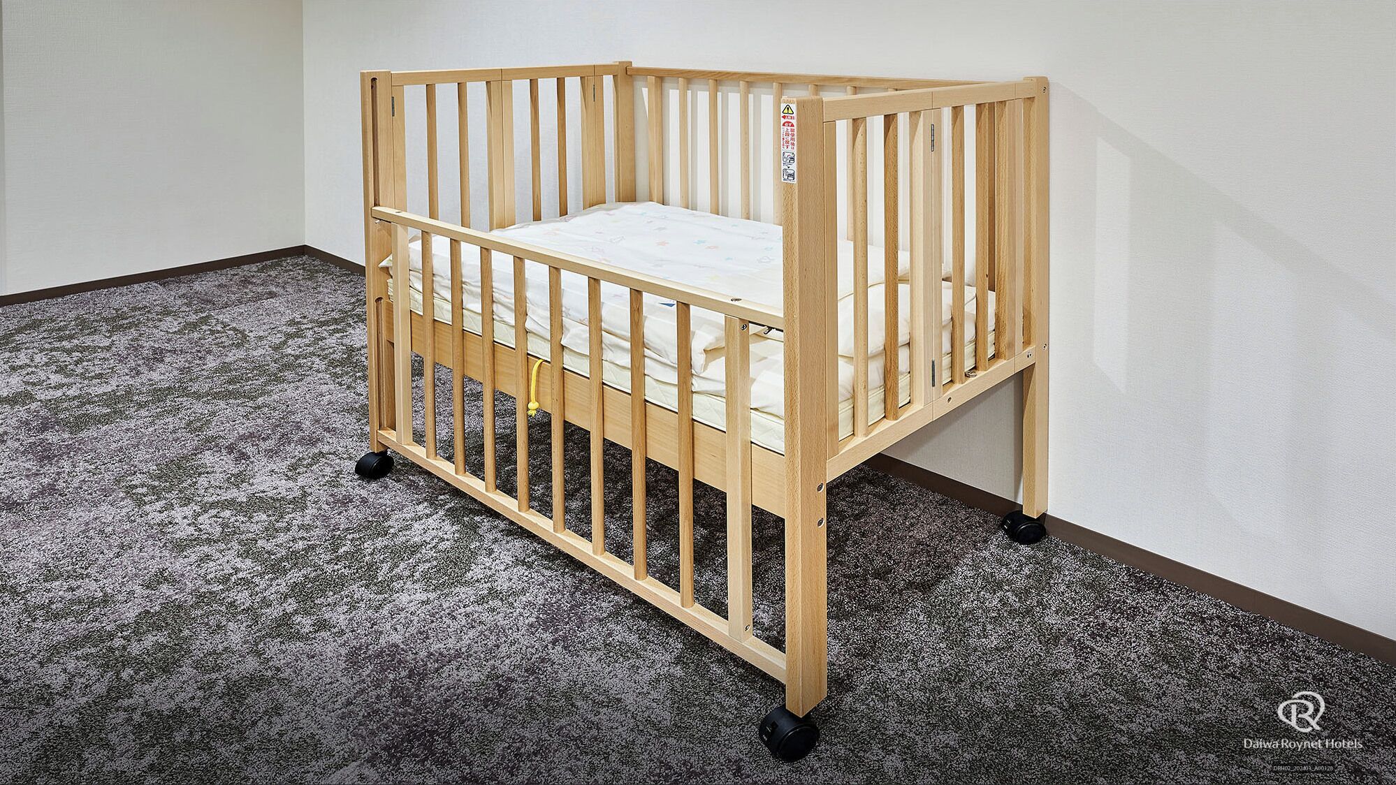 A wooden crib with a mattress, wheels, and a safety guard, placed on a carpeted floor in a room with white walls.