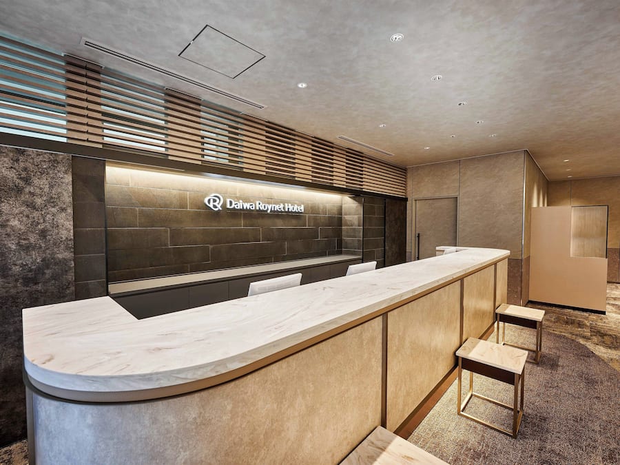 A modern reception area with a marble counter, a logo for Daiwa Roynet Hotel, and a seating area with stools.
