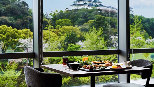 Une table à manger dressée avec des aliments pour le petit-déjeuner et un verre de jus d’orange, donnant sur un château et une végétation luxuriante.