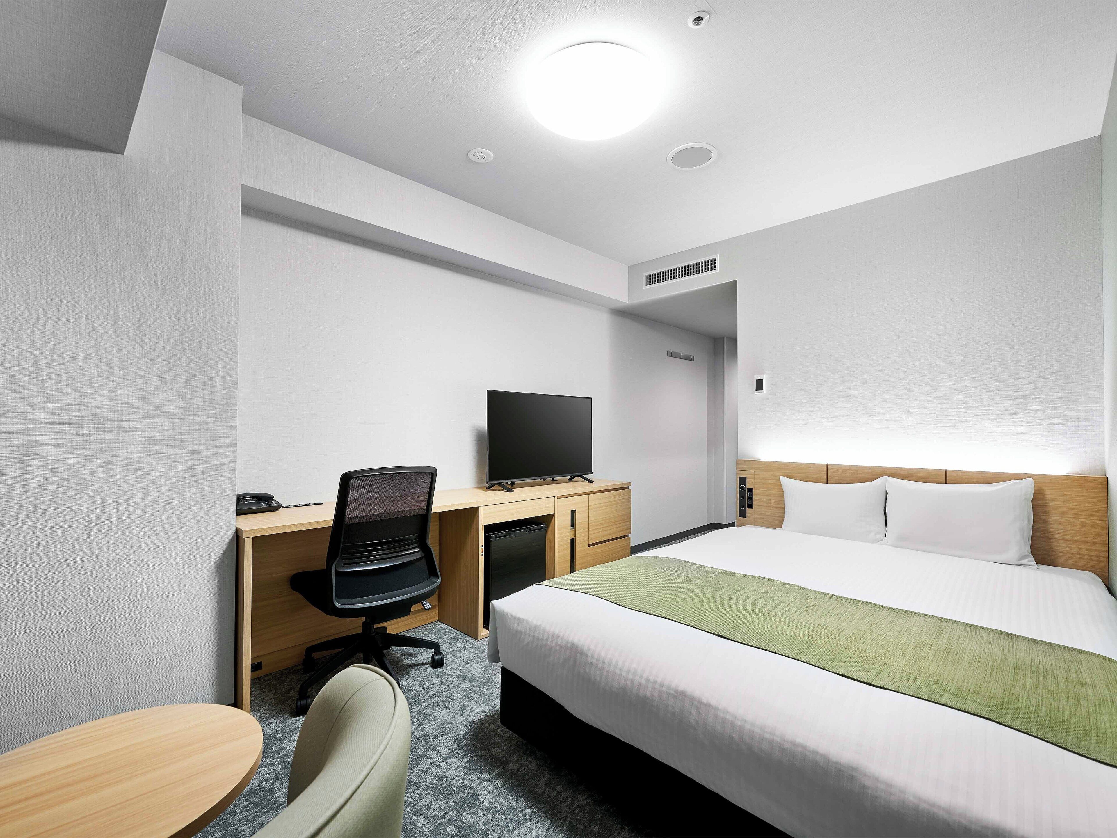 A hotel room with a bed, a desk with a chair, a television, and a small table.