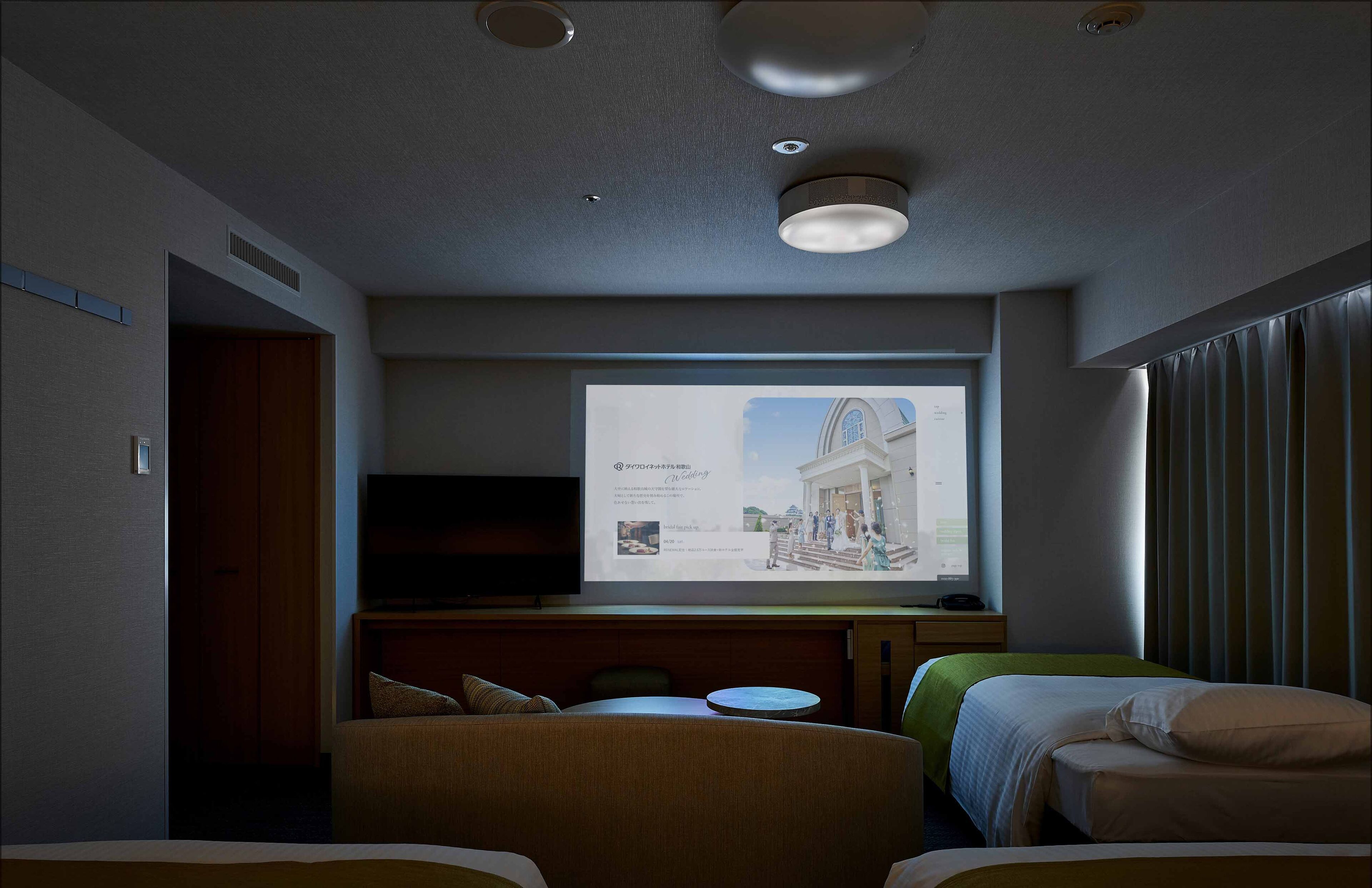 Quarto de hotel com cama, sofá, televisão exibindo uma apresentação e janela com cortinas.