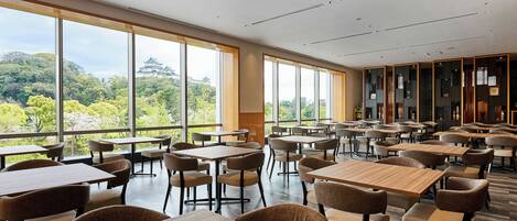 A spacious dining area with large windows offering a view of traditional architecture and greenery. Tables and chairs are arranged neatly.