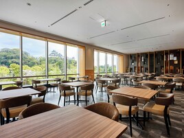 A spacious dining area with large windows offering a view of traditional architecture and greenery. Tables and chairs are arranged neatly.