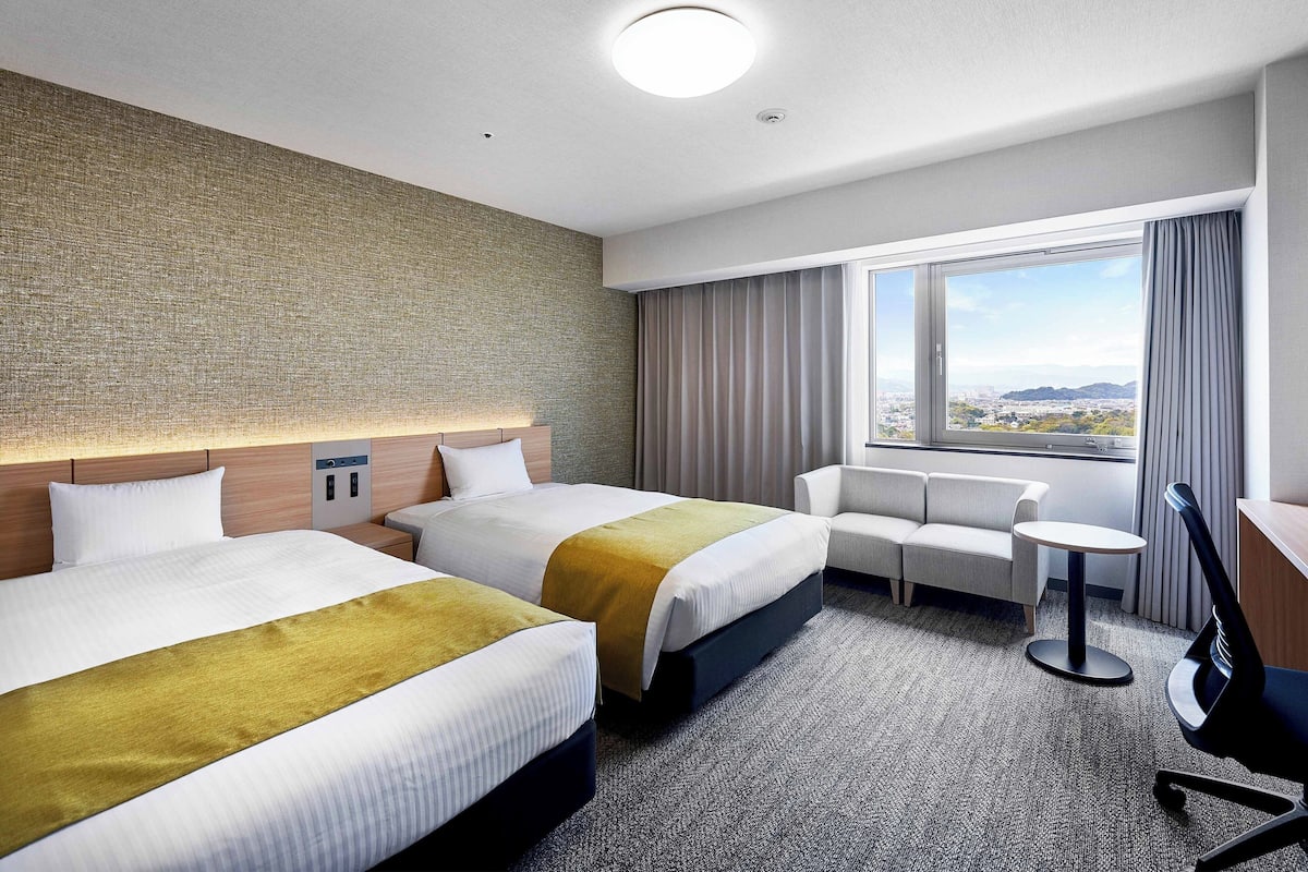 Ein Hotelzimmer mit zwei Betten, einem Sofa, einem Schreibtisch und Blick auf die Stadt.