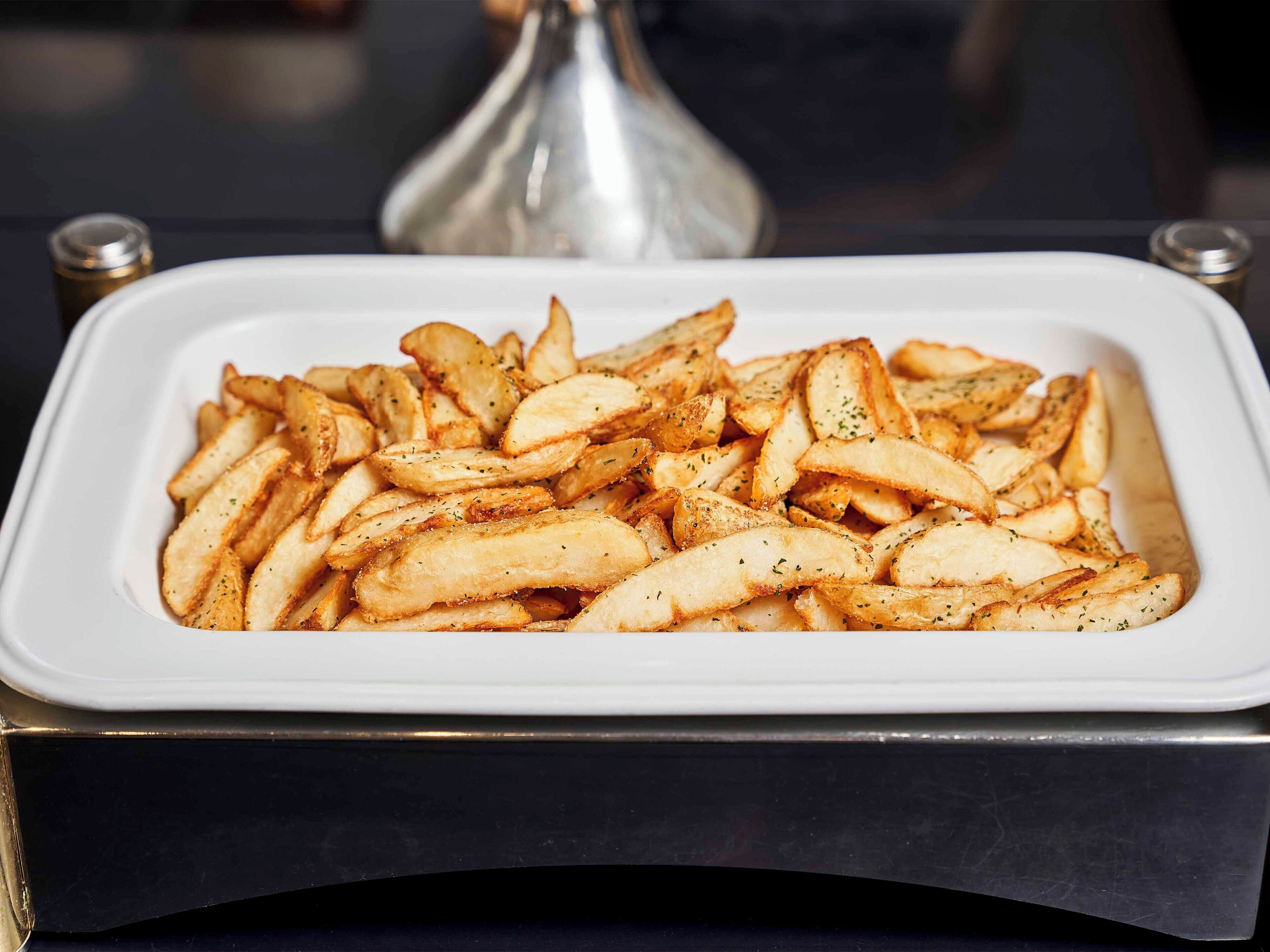 A white rectangular dish filled with golden-brown potato wedges, seasoned with herbs.
