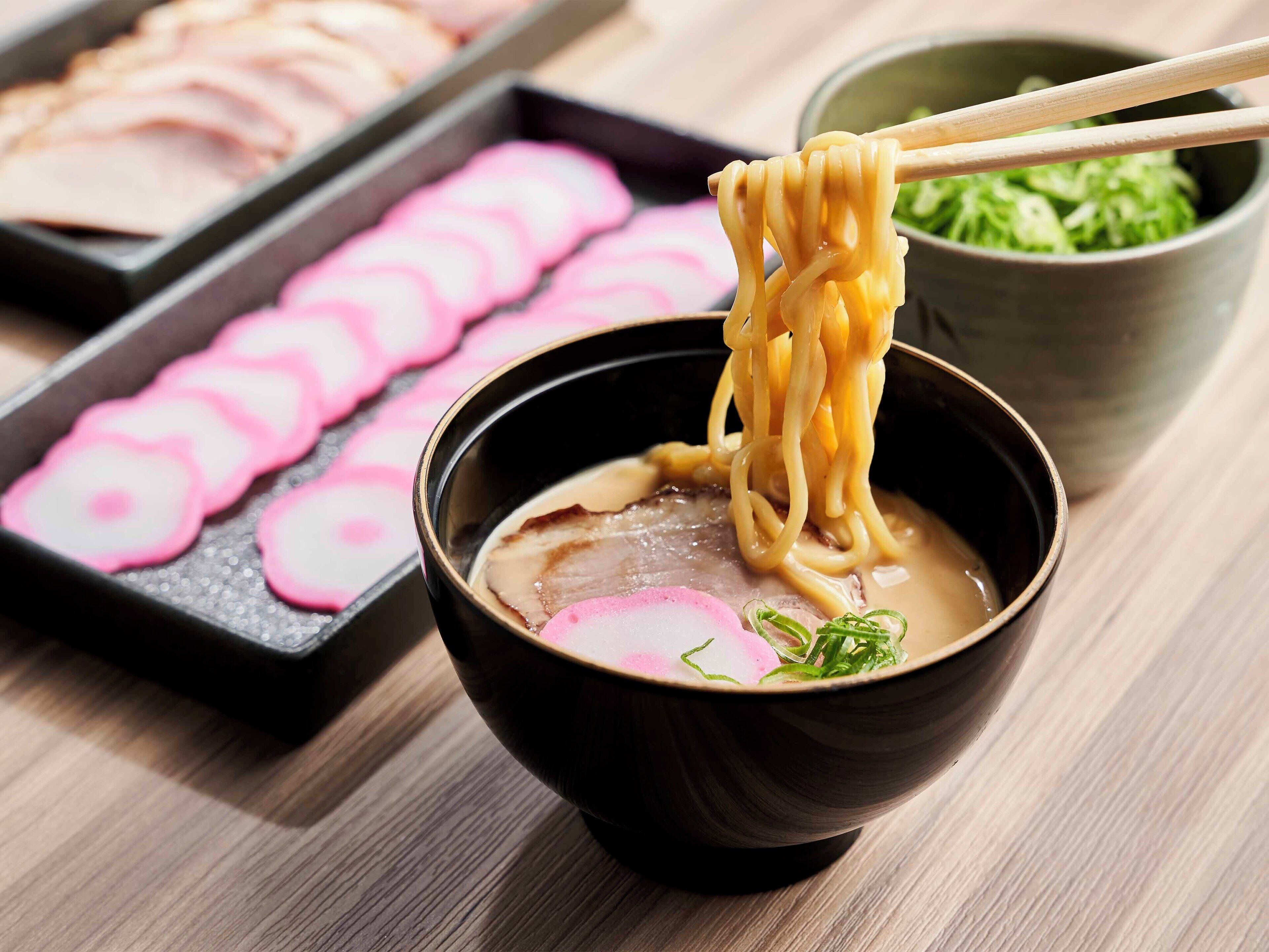 A bowl of ramen noodles being lifted with chopsticks, accompanied by a plate of sliced radishes and a small bowl of green vegetables.