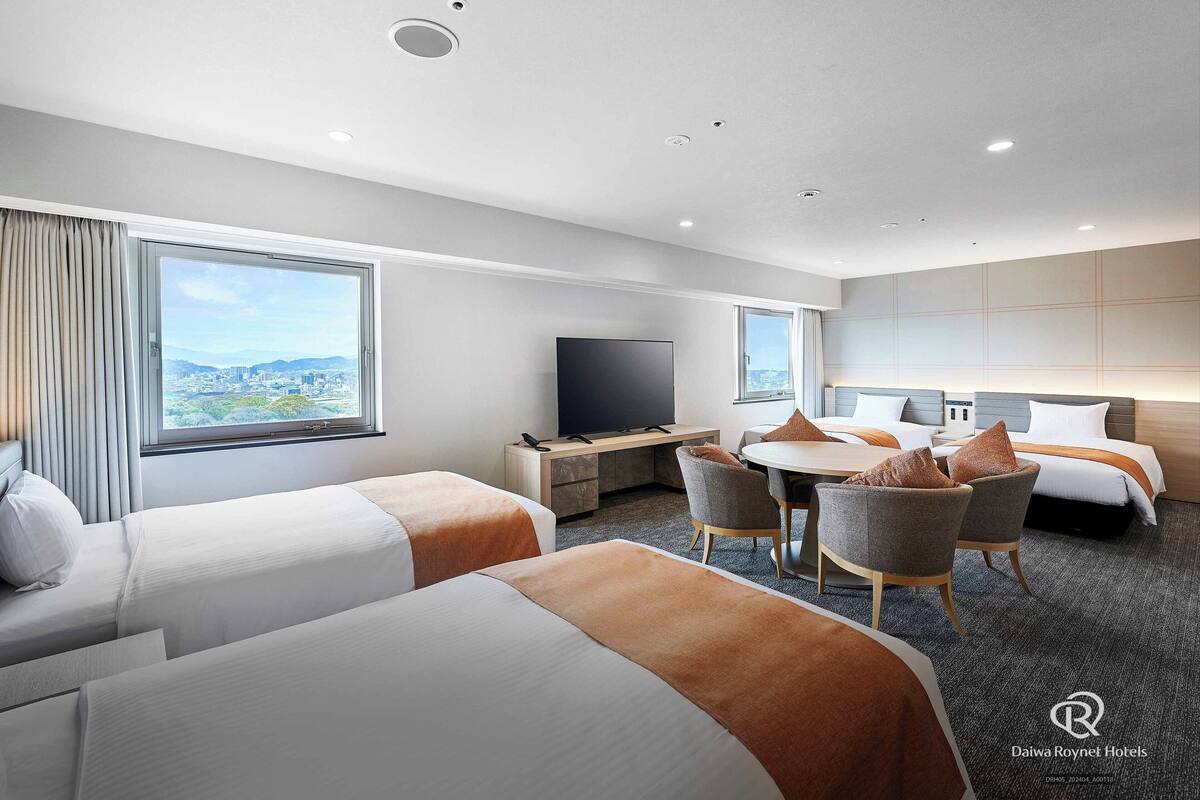 Ein Hotelzimmer mit zwei Betten, einem Esstisch mit Stühlen, einem Fernseher und großen Fenstern mit Blick auf die Stadt.