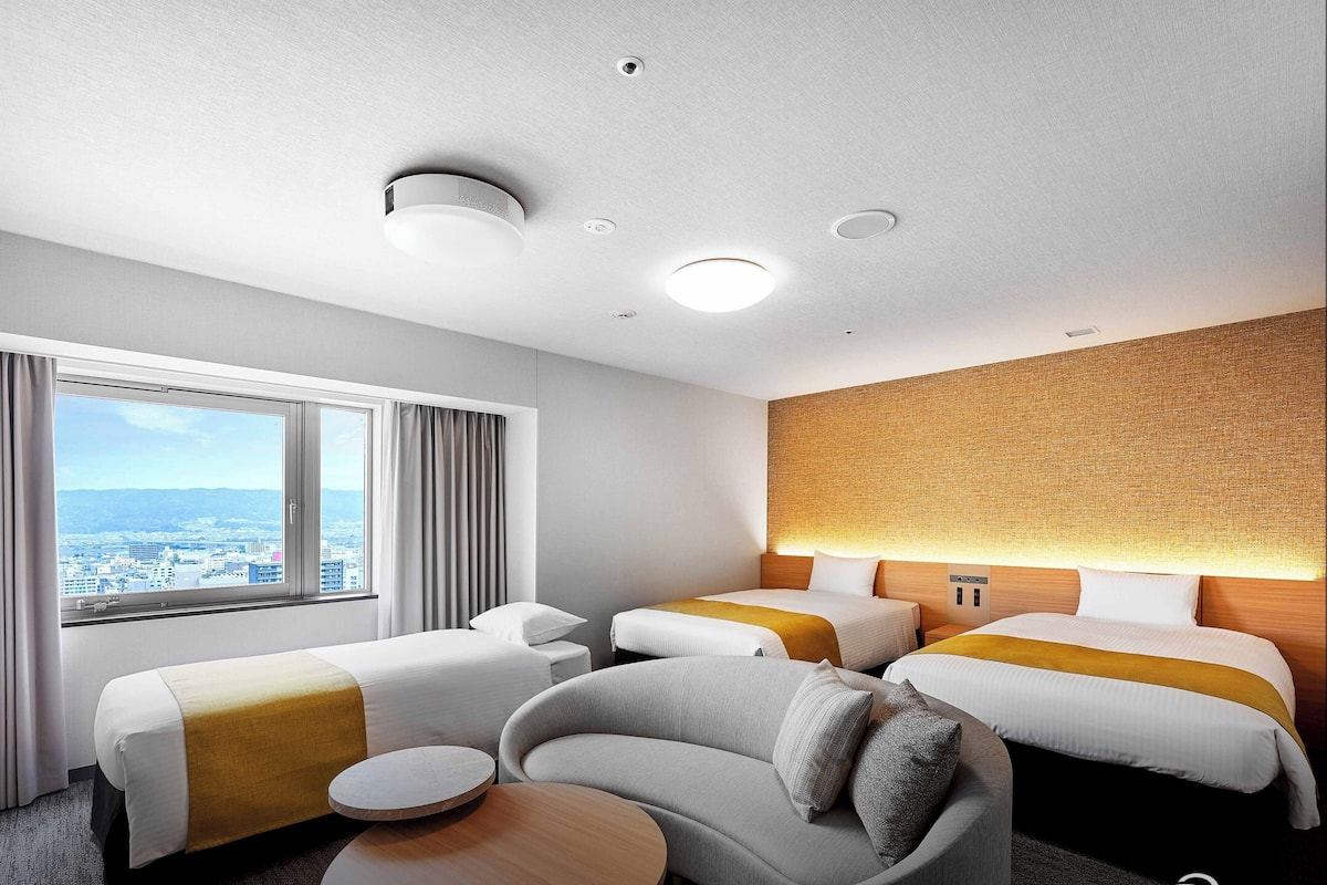 Ein Hotelzimmer mit zwei Betten, einem Sofa, einem kleinen Tisch und einem großen Fenster mit Blick auf die Stadt.