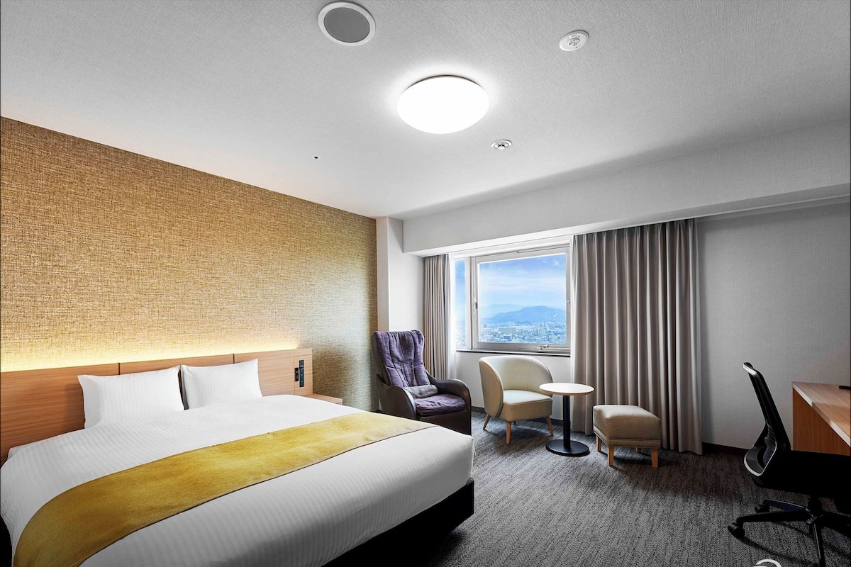 Ein Hotelzimmer mit einem großen Bett, einem Massagesessel, einem Schreibtisch und Blick auf die Stadt durch das Fenster.