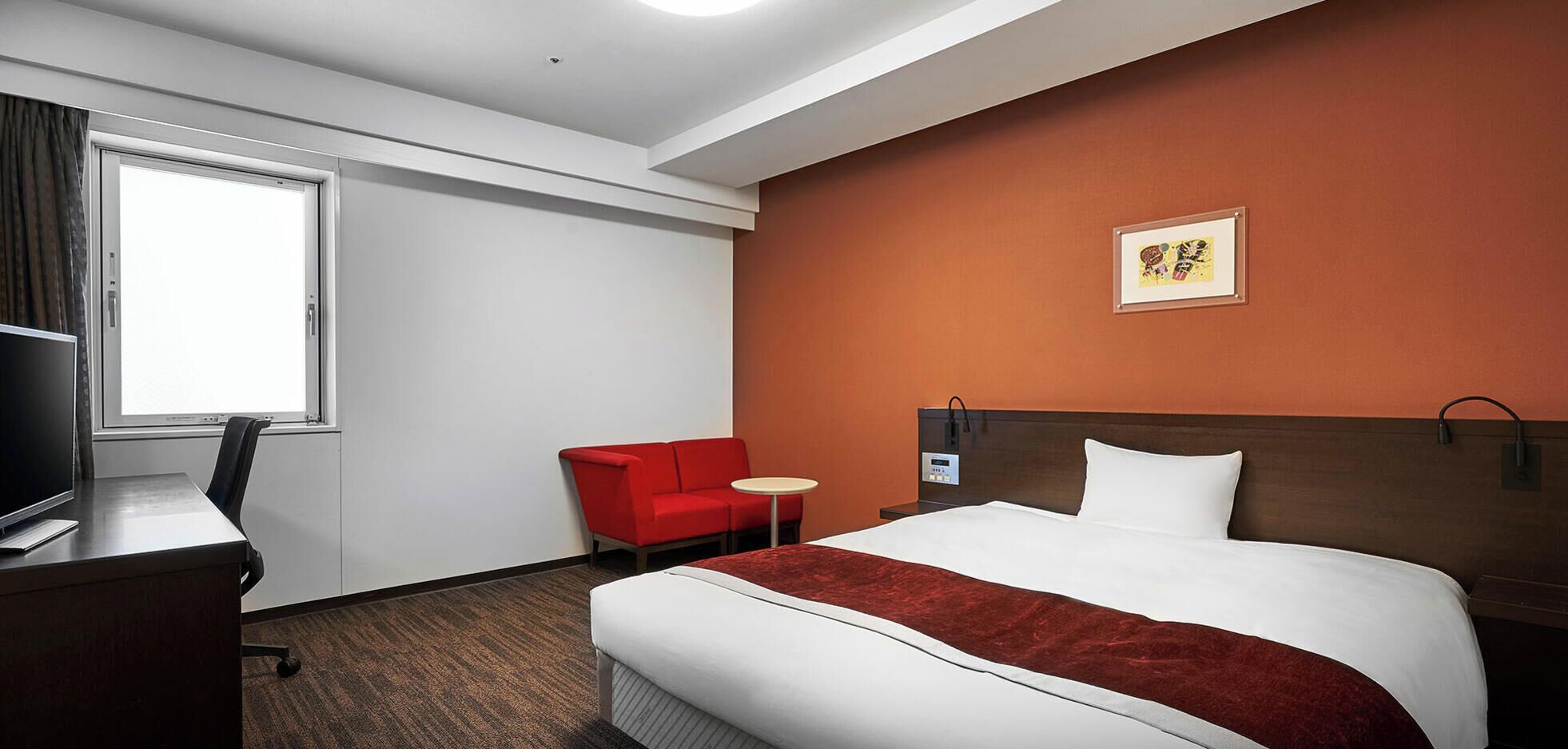 Hotelkamer met een bed, bureau met computer, rode fauteuil, klein rond tafeltje en een tv aan de muur.