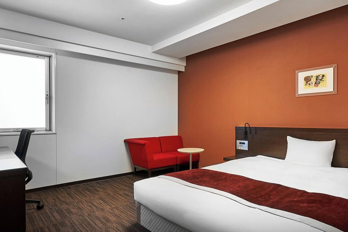 Une chambre d’hôtel avec un lit, un bureau équipé d’un ordinateur, un fauteuil rouge, une petite table ronde et un téléviseur fixé au mur.