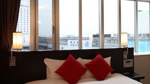 Habitación de hotel con cama, almohadas rojas, mesita de noche, dos lámparas y ventana con vista a la ciudad.