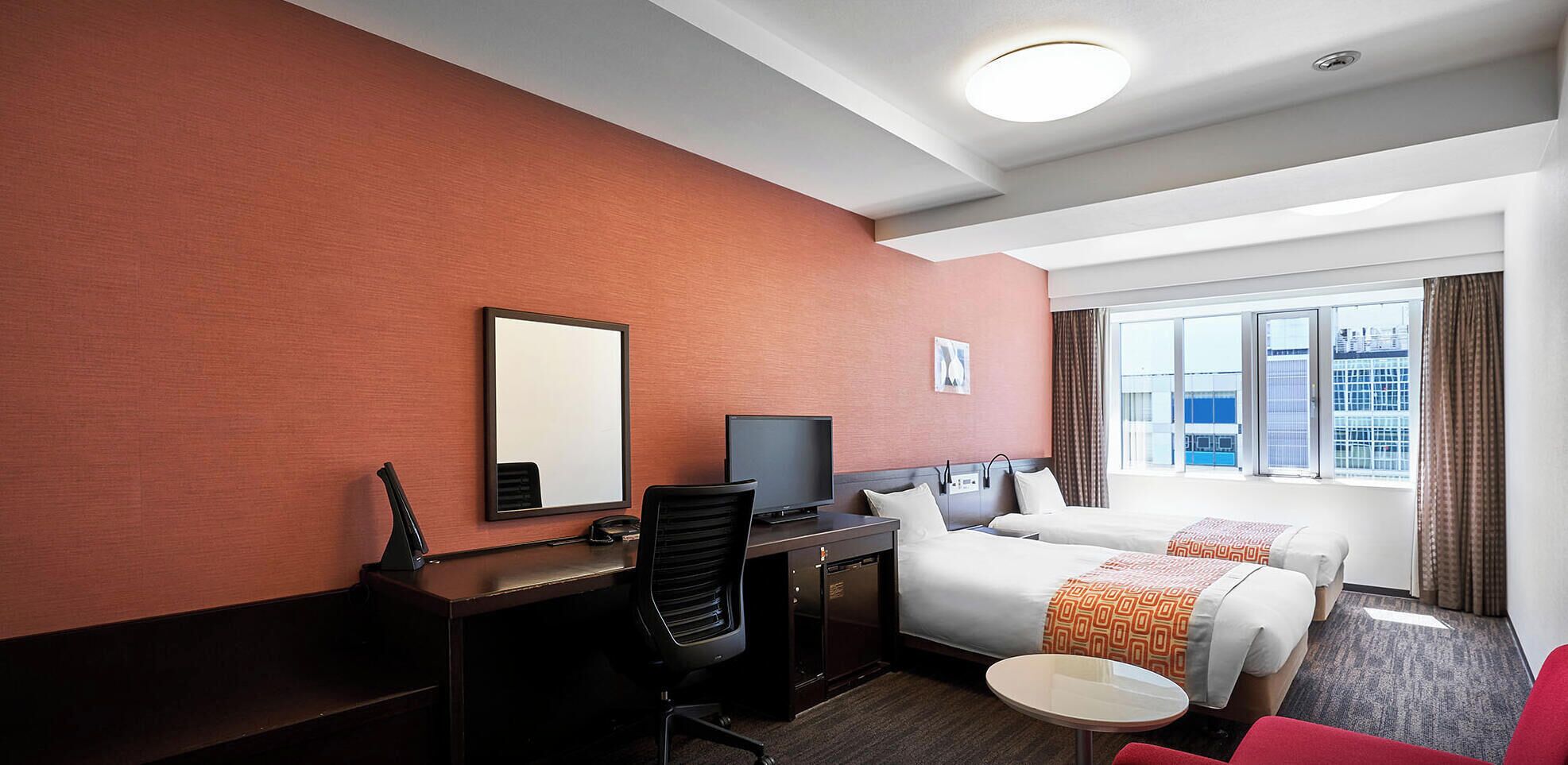Hotelkamer met een bureau, stoel, televisie, twee bedden, een kleine tafel en een raam met uitzicht op de stad.