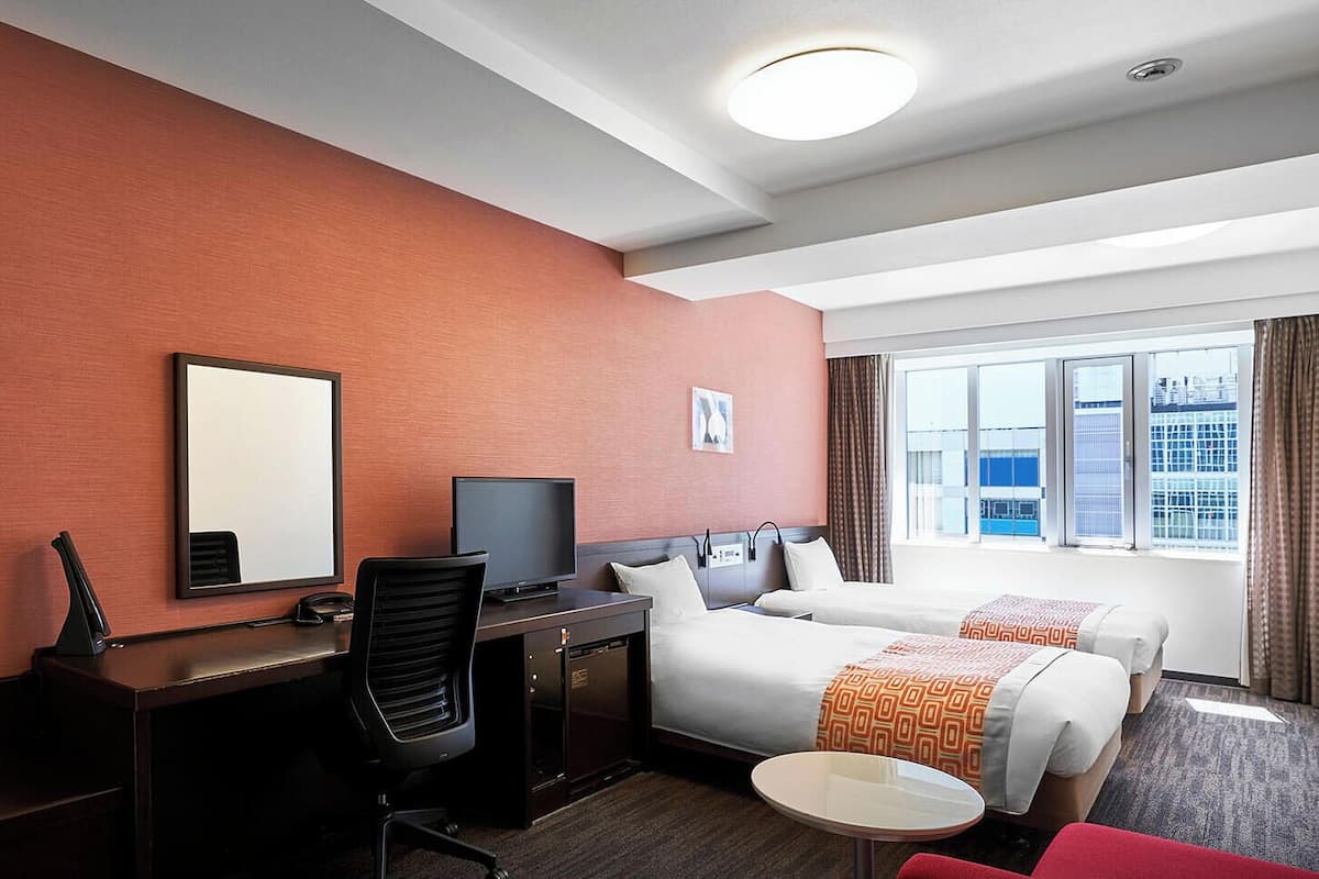 Une chambre d’hôtel équipée d’un bureau, d’une chaise, d’une télévision, de deux lits, d’une petite table et d’une fenêtre donnant sur la ville.