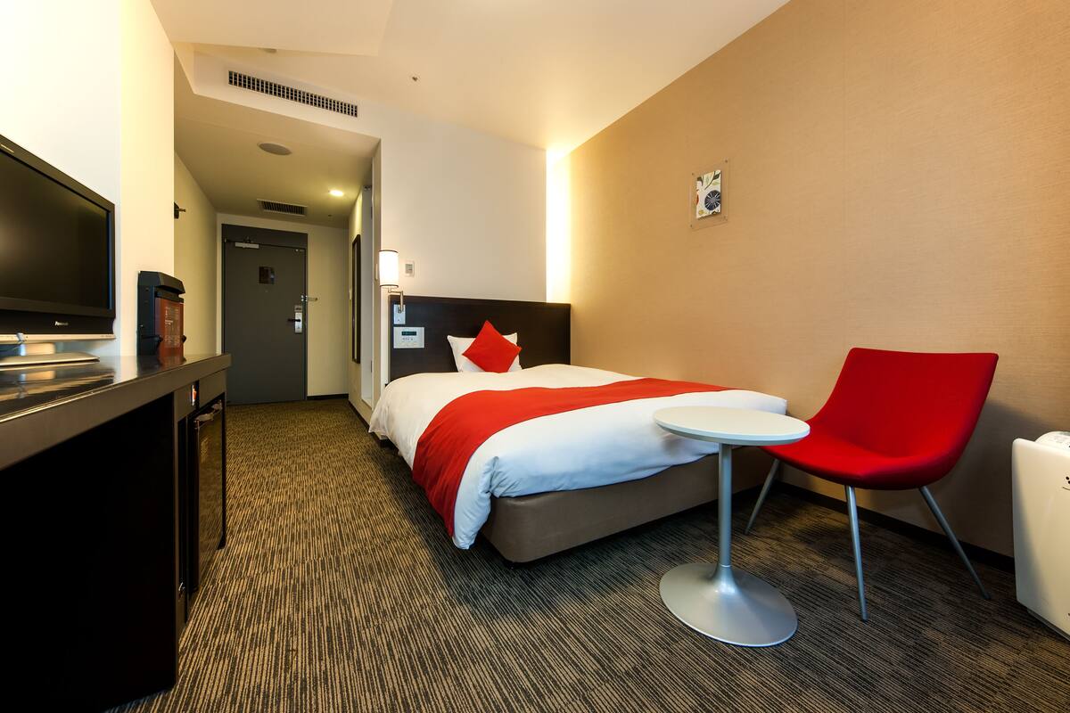 Une chambre d’hôtel avec un lit, une chaise rouge, une petite table, un téléviseur et un mini-bar.
