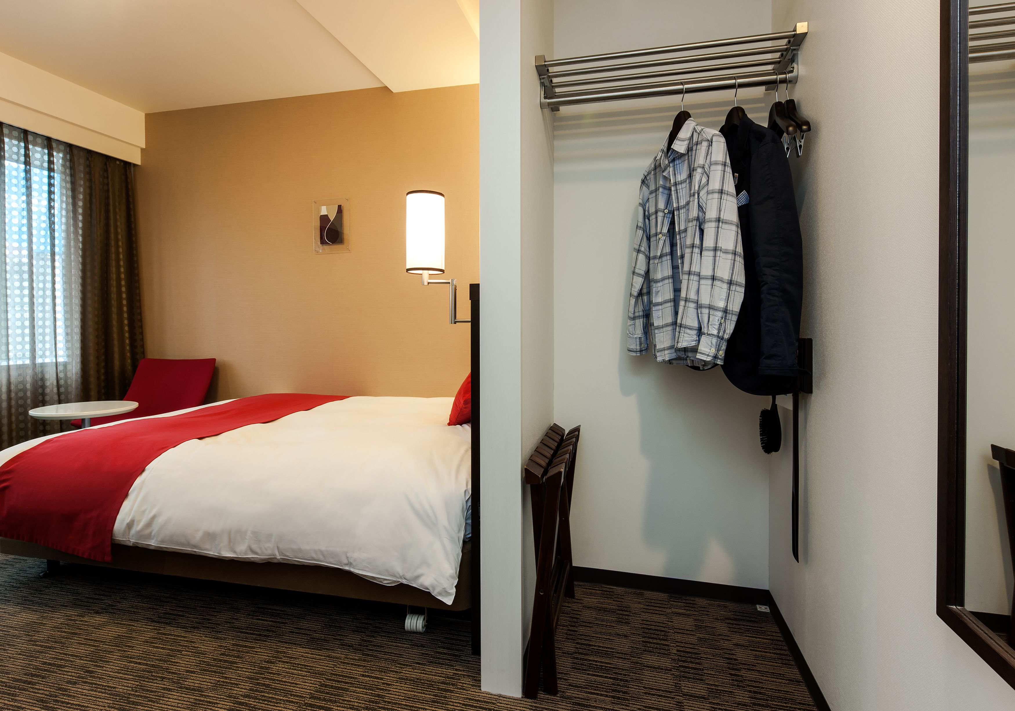 A hotel room with a bed, a small table, a mirror, and a coat rack with hanging clothes.