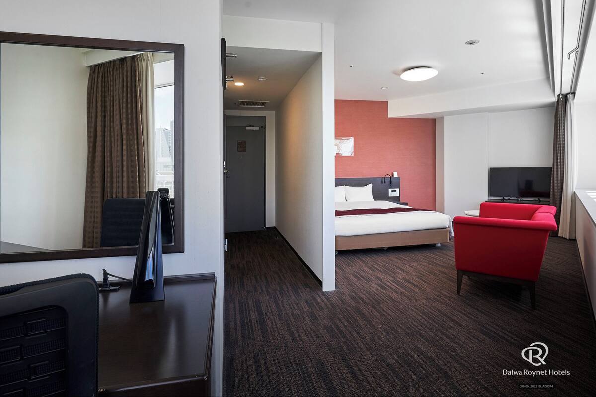 Une chambre d’hôtel avec un lit, un fauteuil rouge, une télévision et un bureau avec un ordinateur.