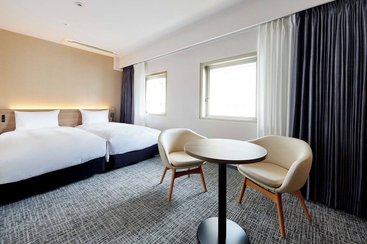 Una habitación de hotel con una cama, dos sillas, una mesita y dos ventanas.