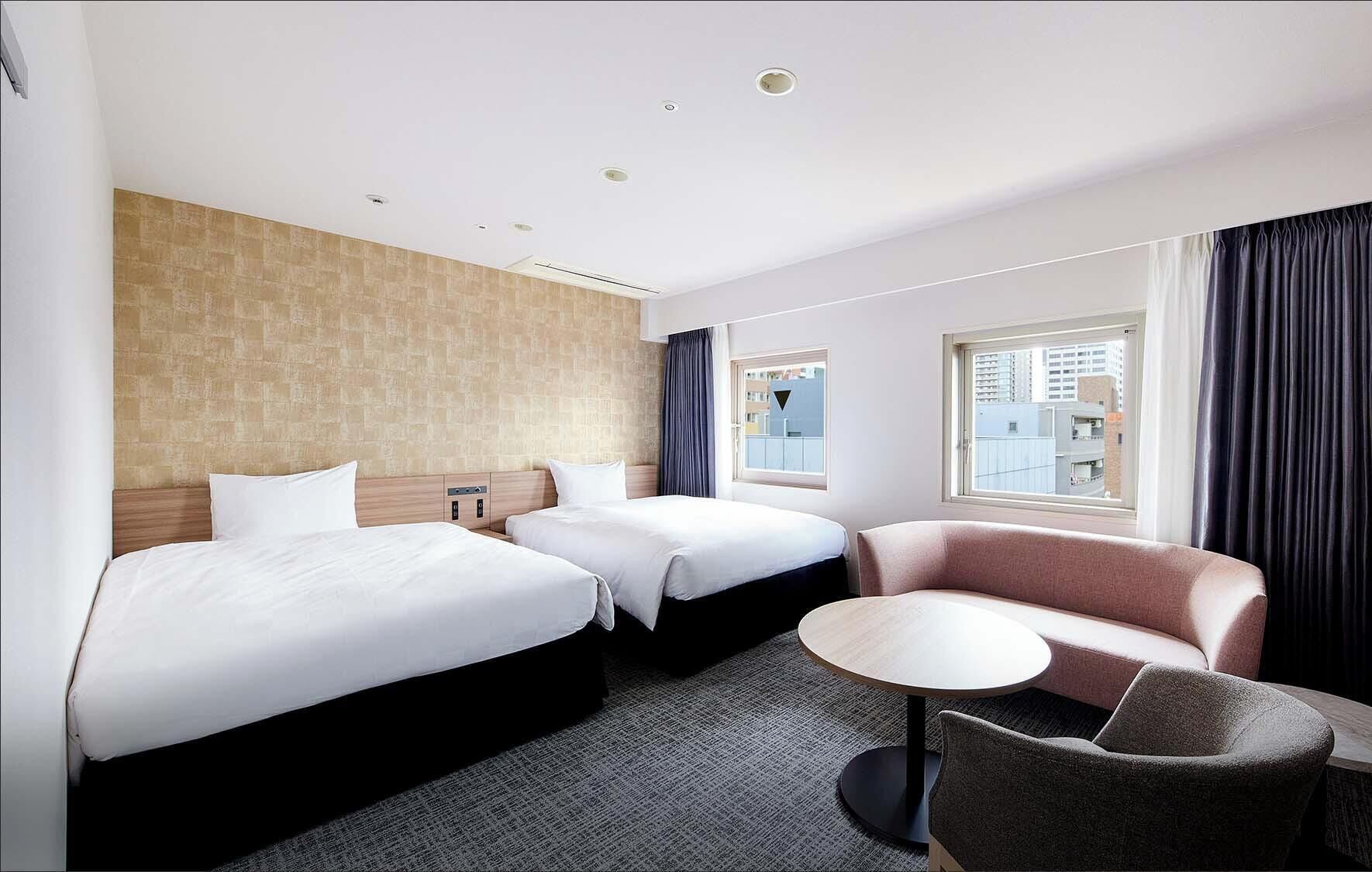 Habitación de hotel con dos camas, una mesita redonda, un sofá y vista a la ciudad por la ventana.