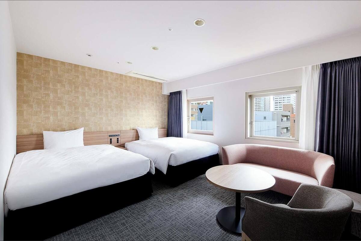 Habitación de hotel con dos camas, una mesita redonda, un sofá y vista a la ciudad por la ventana.