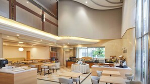 A spacious hotel dining area with multiple seating options, a central counter with a coffee machine, and large windows providing natural light.