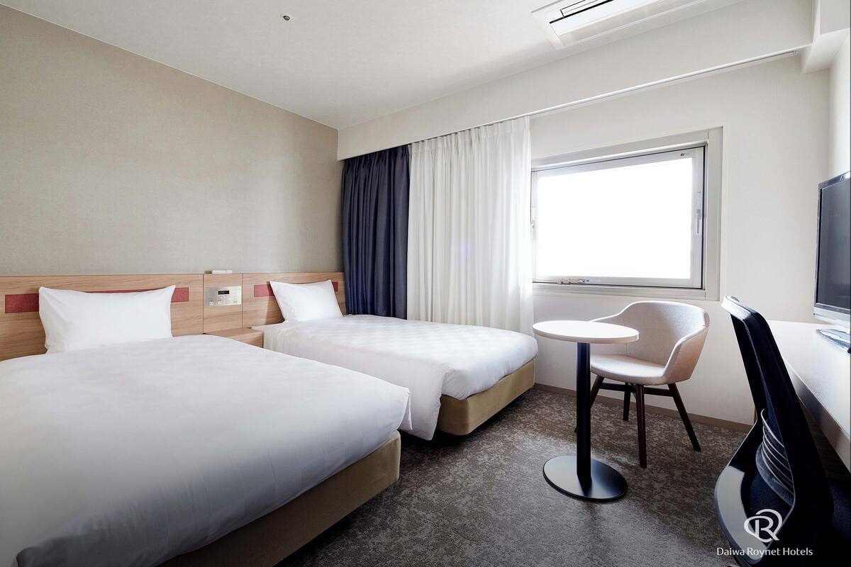 Habitación de hotel con dos camas, una mesita redonda pequeña, una silla, un televisor y un ventanal con cortinas.