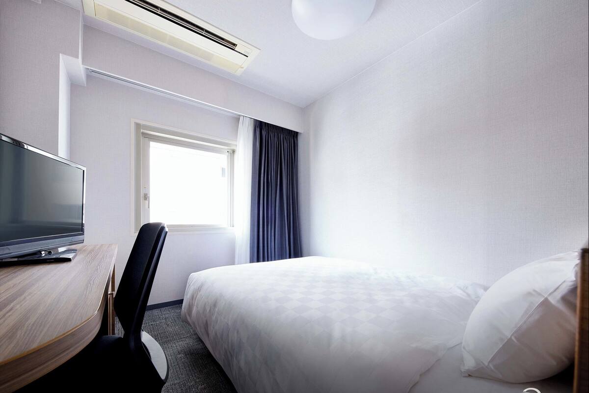 Habitación de hotel con cama, escritorio con televisor, silla, ventana con cortinas e unidad de aire acondicionado.