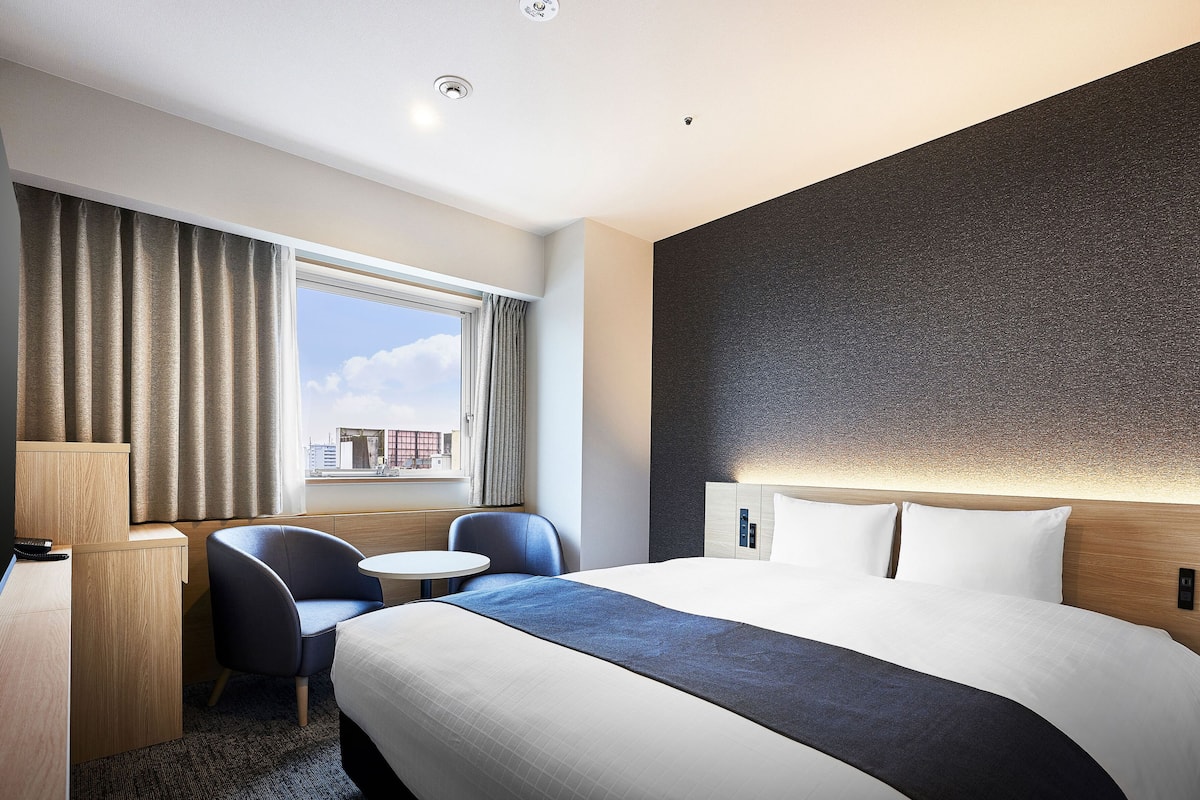 Een hotelkamer met een groot bed, een bureau, twee stoelen, een kleine tafel en een raam met gordijnen.