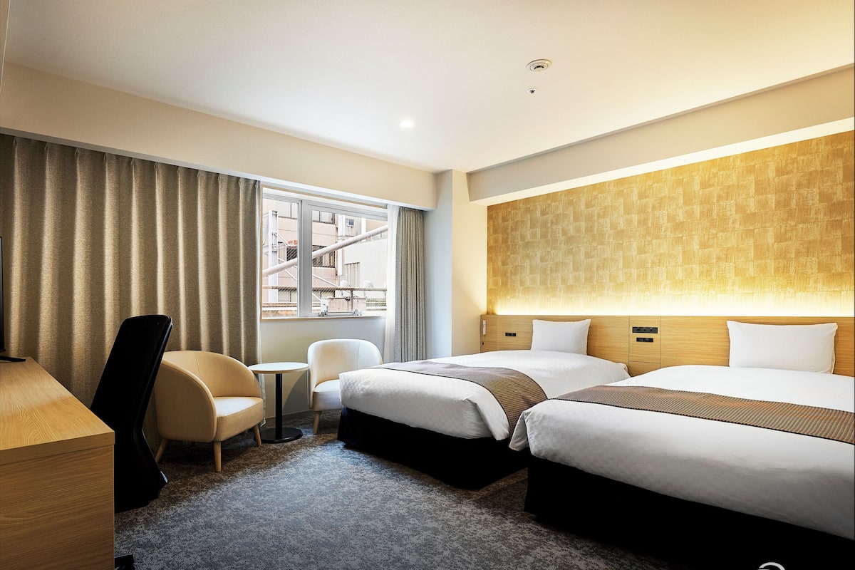 Een hotelkamer met twee bedden, een bureau, een stoel en uitzicht op de stad door het raam.