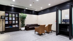 A lobby with a black and white tiled floor, a brown leather sofa, two brown armchairs, a large potted plant, a black bookshelf with magazines, and a computer monitor on a black stand. - Daiwa Roynet Hotel Kobe Sannomiya (Kobe)