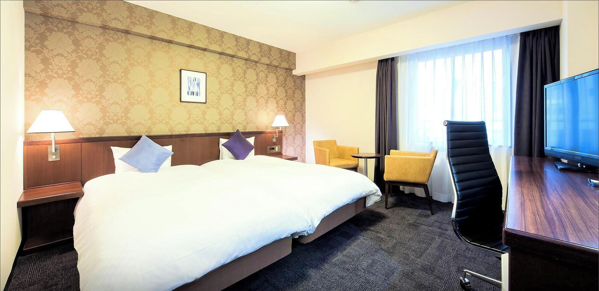 A hotel room with a large bed, two bedside lamps, a desk with a flat-screen TV, a chair, and a window with curtains.