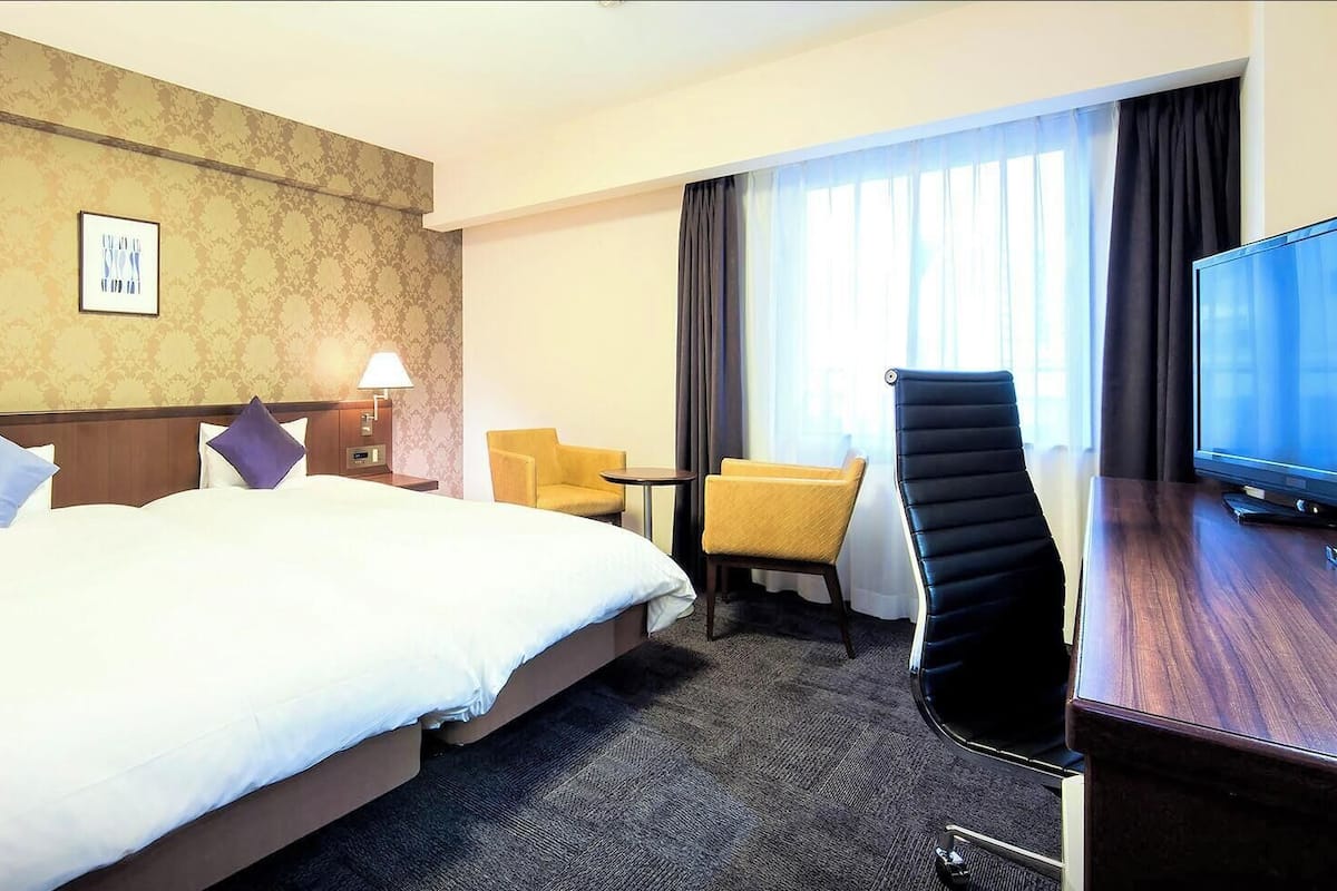 Une chambre d’hôtel avec un grand lit, deux lampes de chevet, un bureau avec un téléviseur à écran plat, une chaise et une fenêtre avec des rideaux.