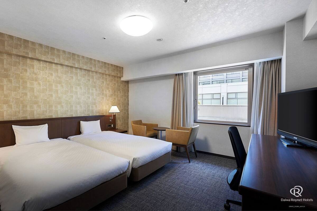Une chambre d’hôtel avec deux lits, un bureau, une chaise, une télévision et une fenêtre avec des rideaux.