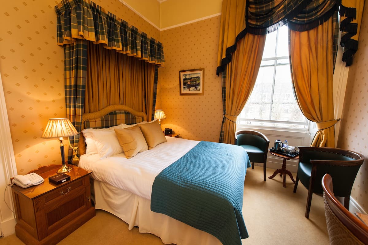 A hotel room with a bed, bedside lamps, a chair, a small table, and a window with curtains.
