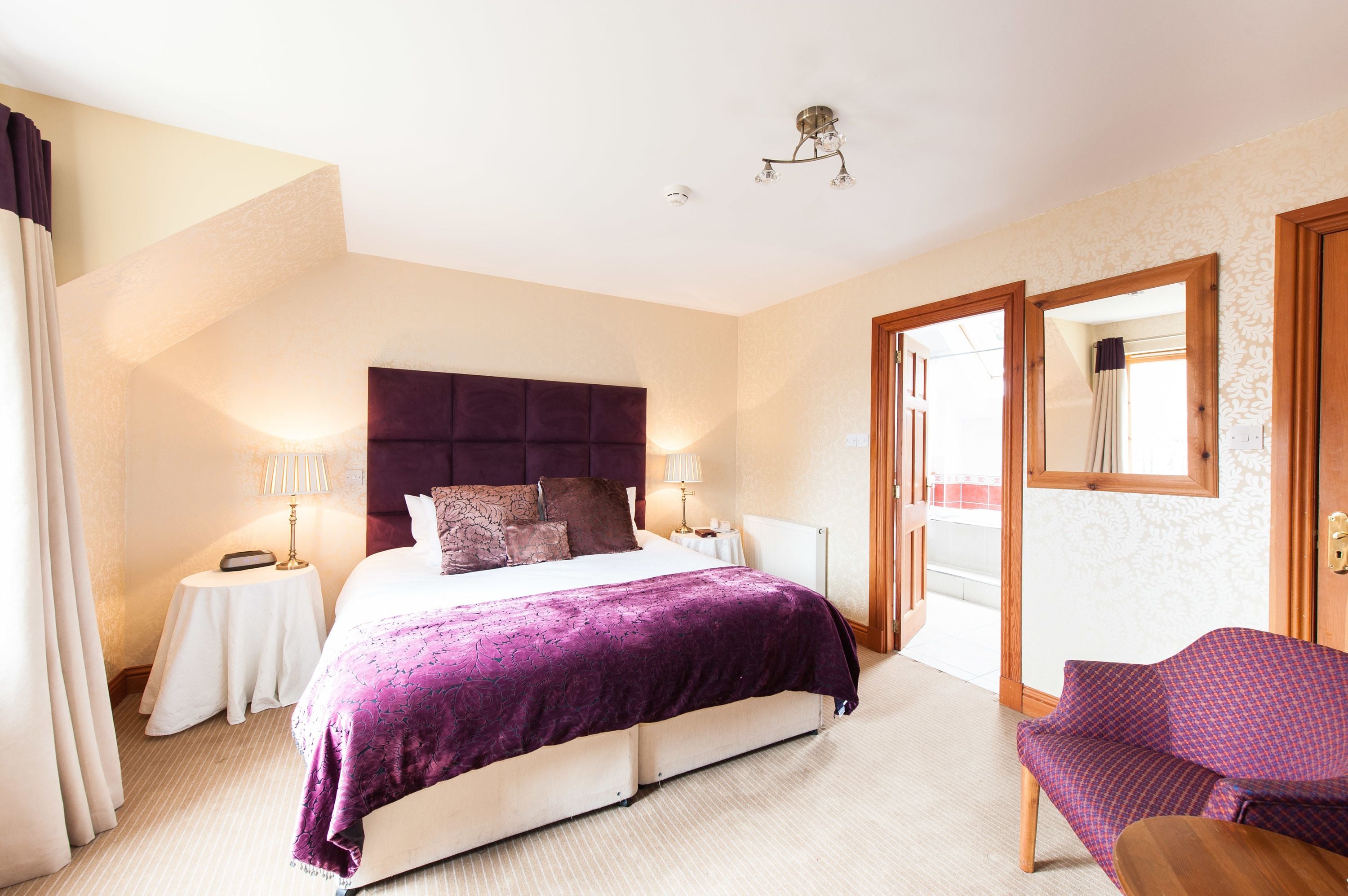 A hotel room with a large bed, a small table with a lamp, a chair, and a large mirror.